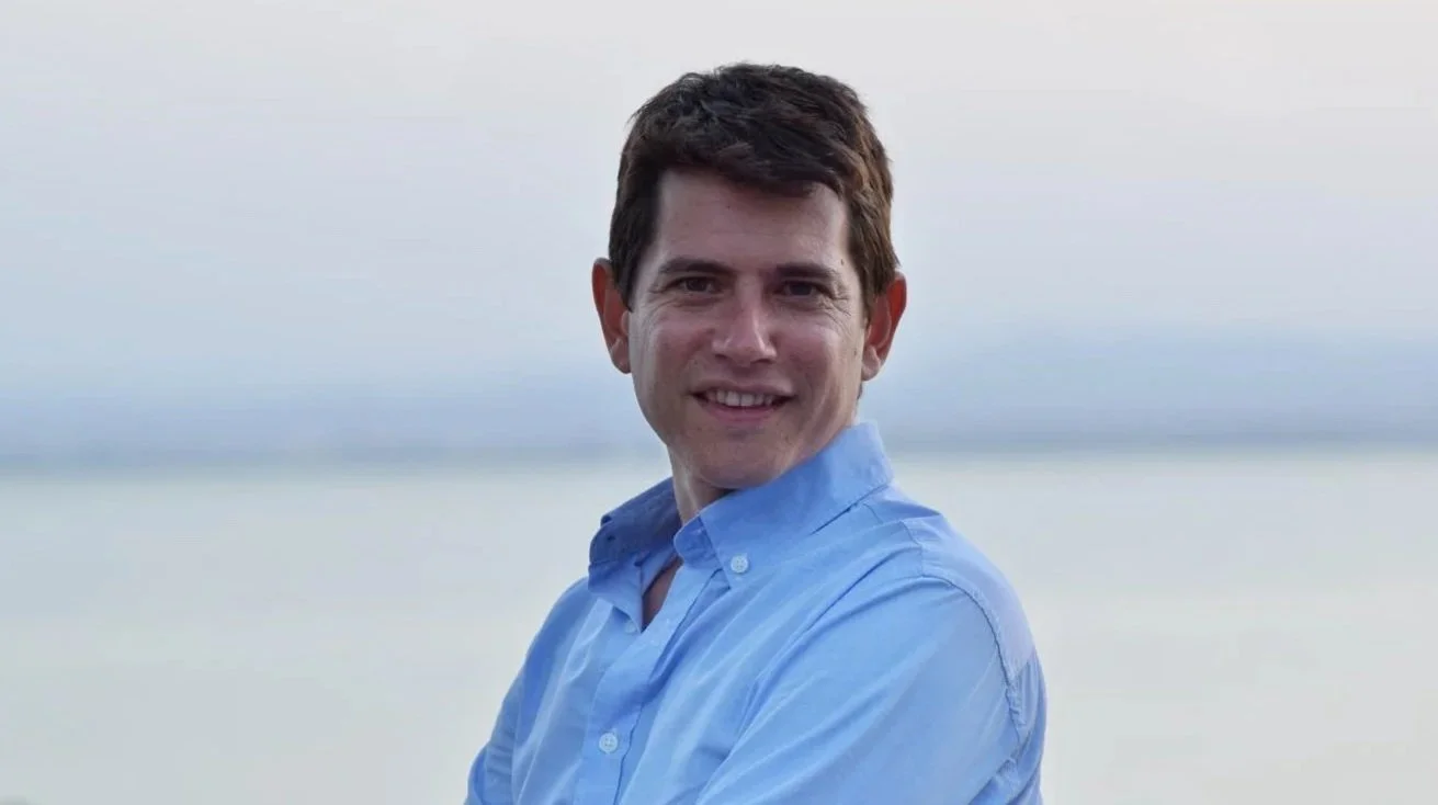 A young man with brown hair wearing a blue button-up shirt, smiling, outdoors near a body of water with a cloudy sky in the background.