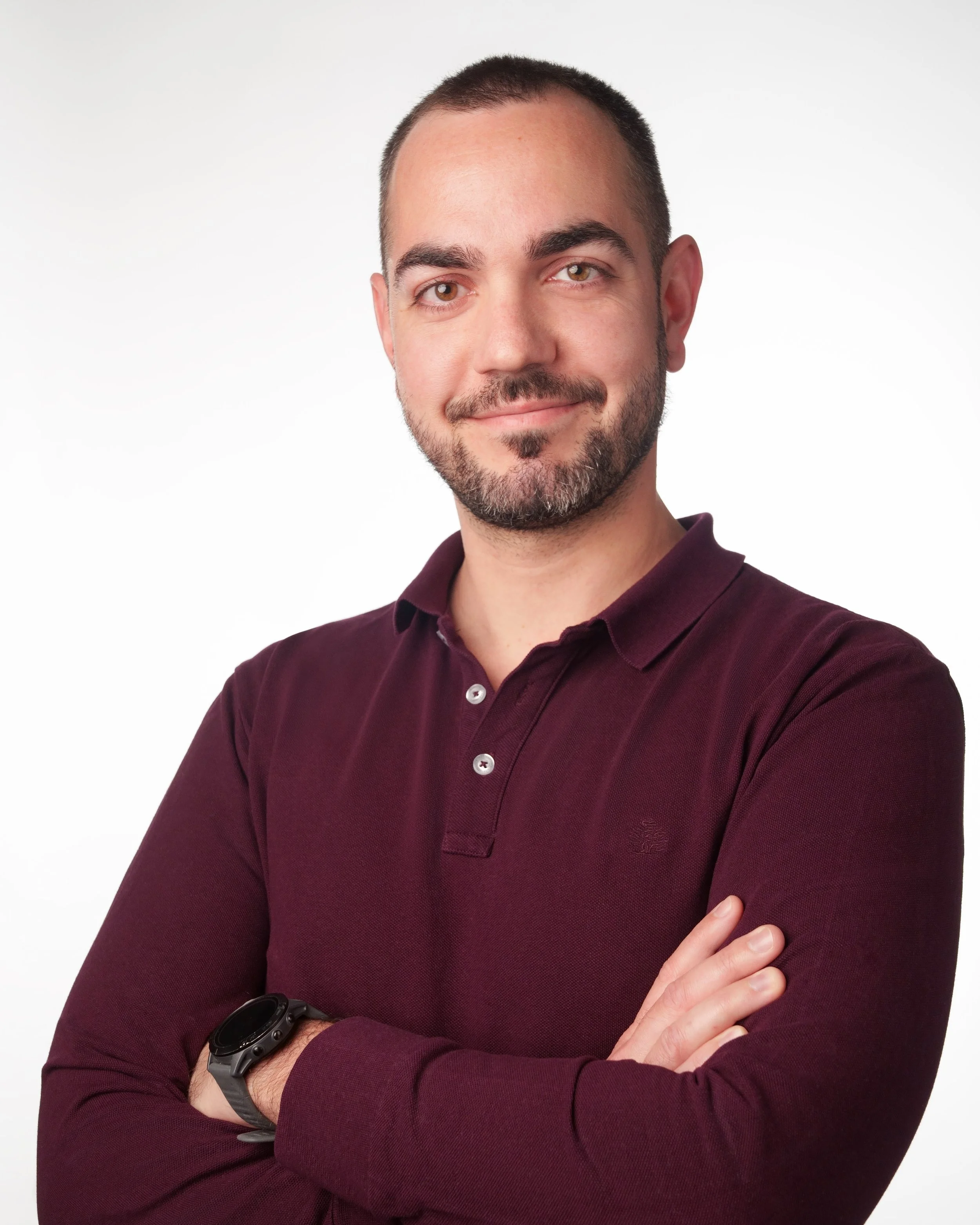 Porträt eines Mannes mit dunklem Bart, kurzhaarig, trägt einen dunkelroten Polo, steht vor weißem Hintergrund, lächelnd mit verschränkten Armen, trägt eine Smartwatch an seinem linken Handgelenk.