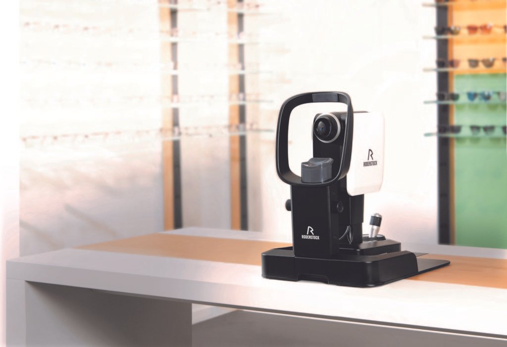 Optischer Scanner auf einem Tisch, mit einem Hintergrund von Regal mit Brillen. Das Gerät ist schwarz und weiß mit einem runden Lesekopf.