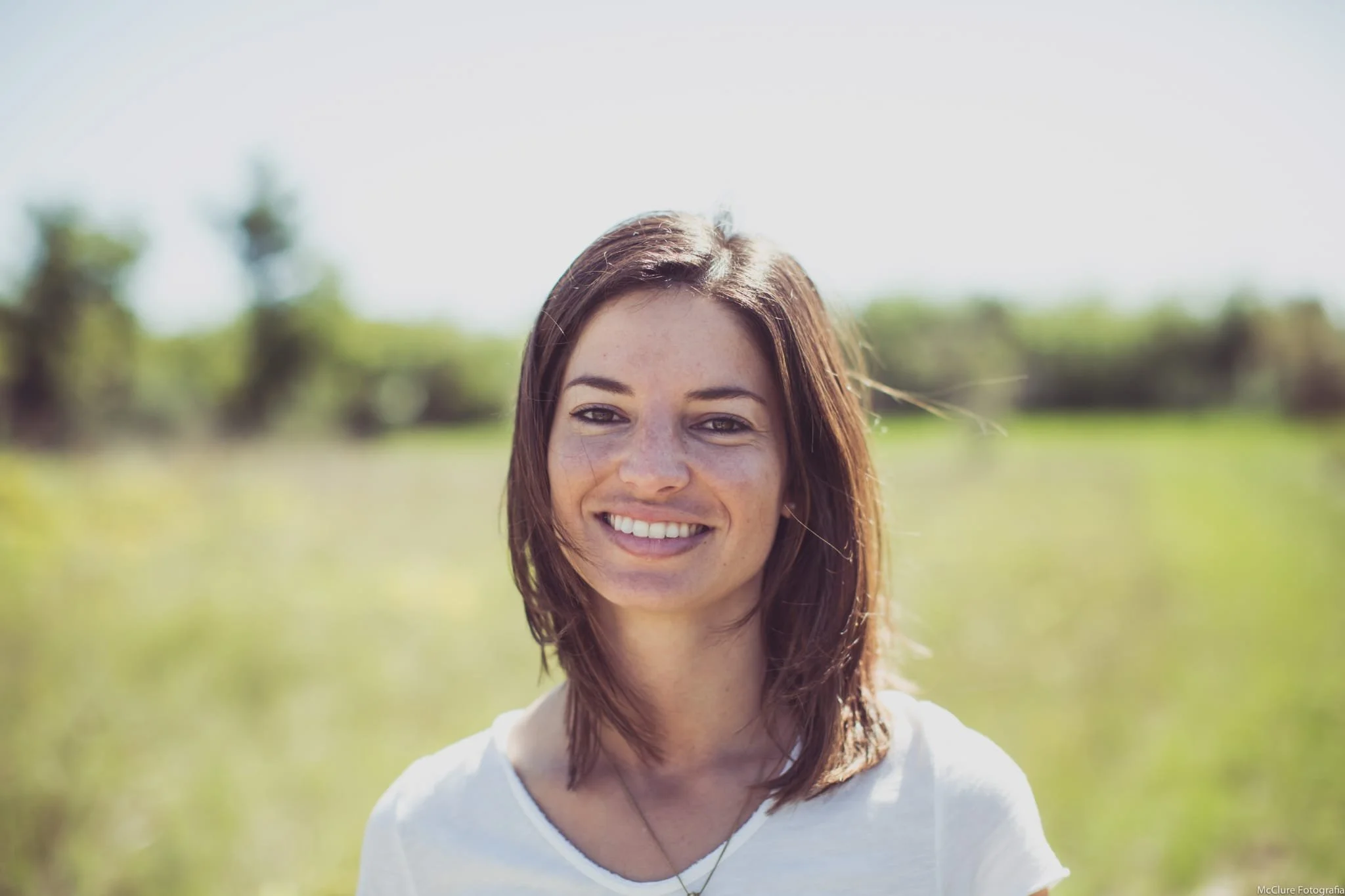 A young woman with shoulder-length brown hair, smiling outdoors in a sunny field with blurred green trees in the background.
