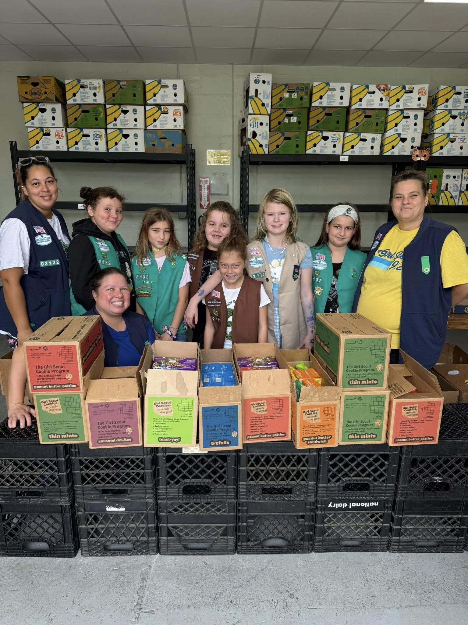 A group of girls in Girl Scout uniforms and two adult leaders standing behind a table filled with boxes of Girl Scout cookies in a warehouse or storage room.