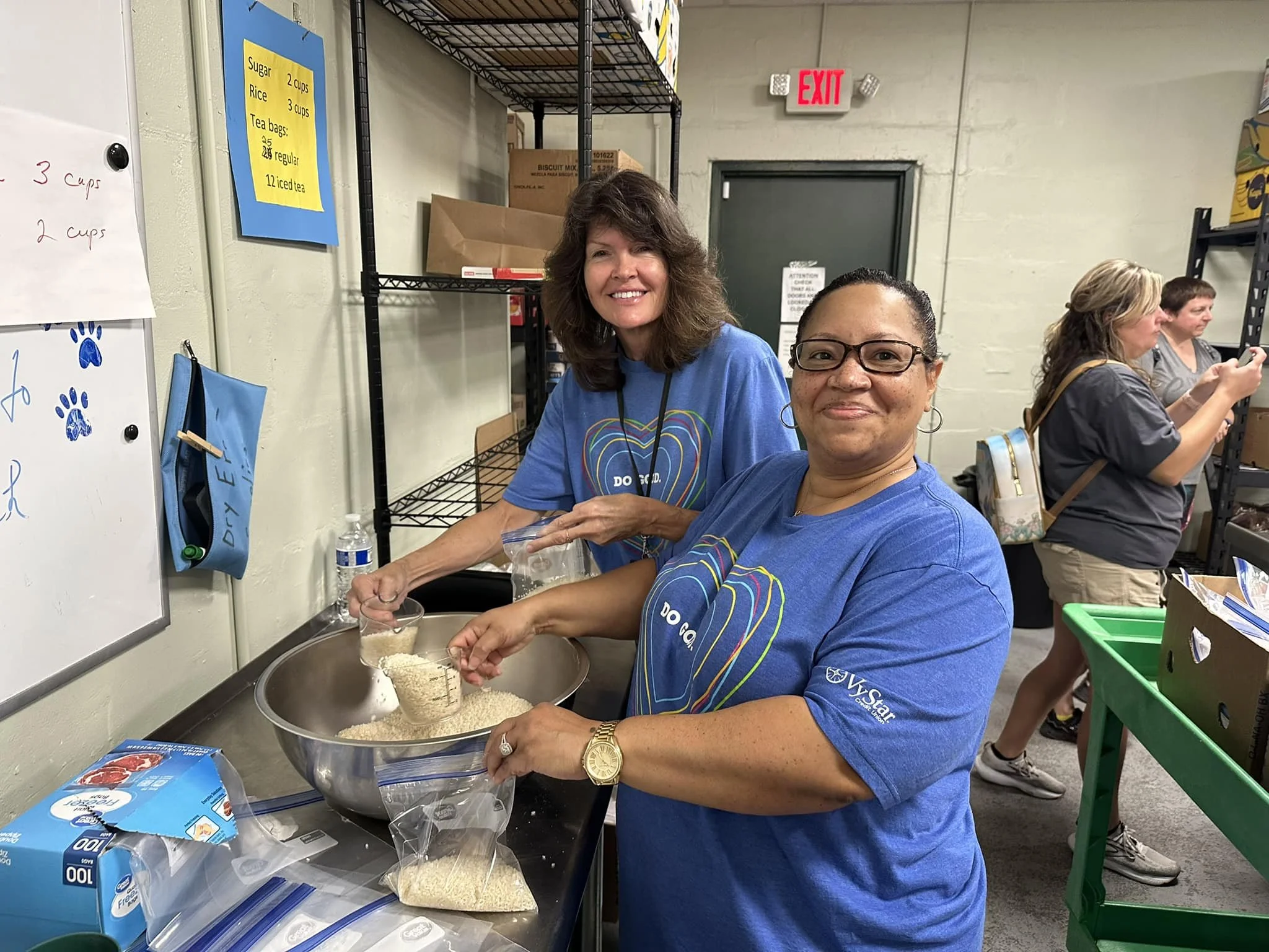 Two women in blue shirts preparing food in a warehouse or kitchen setting. They are smiling, with one pouring rice from a measuring cup into a plastic bag, and various food items and supplies are visible around them.