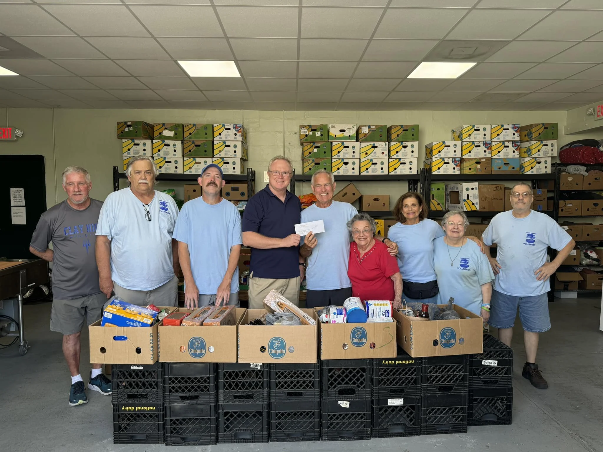 Group of people standing behind boxes filled with food donations in a room with shelves stocked with boxes.