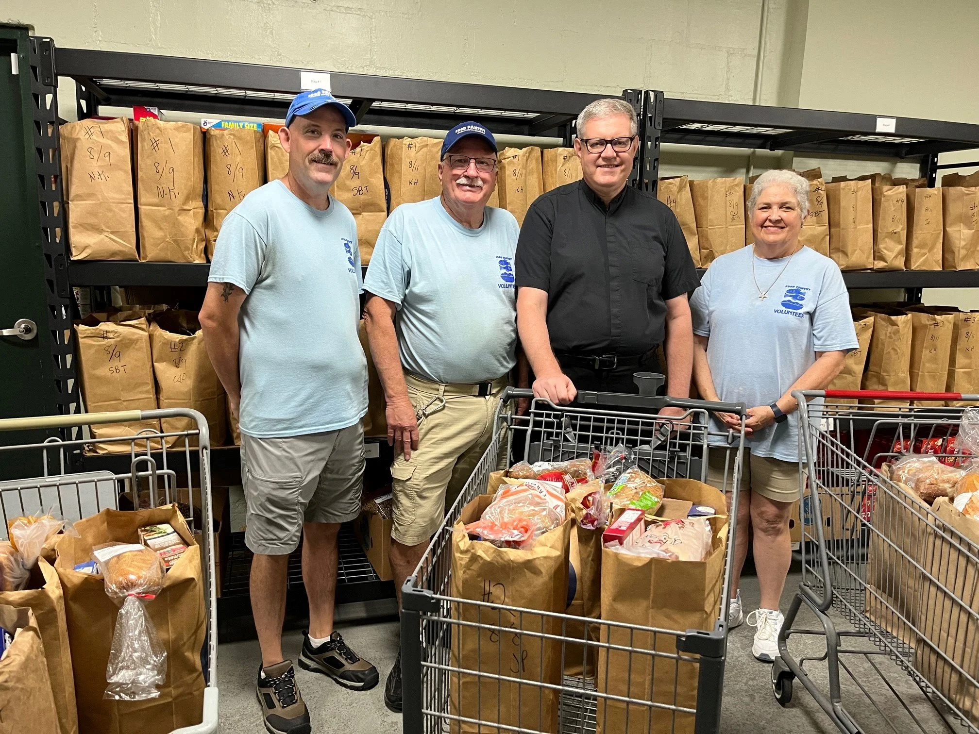 Four people standing behind shopping carts filled with groceries at a food bank, with shelves filled with paper bags in the background.