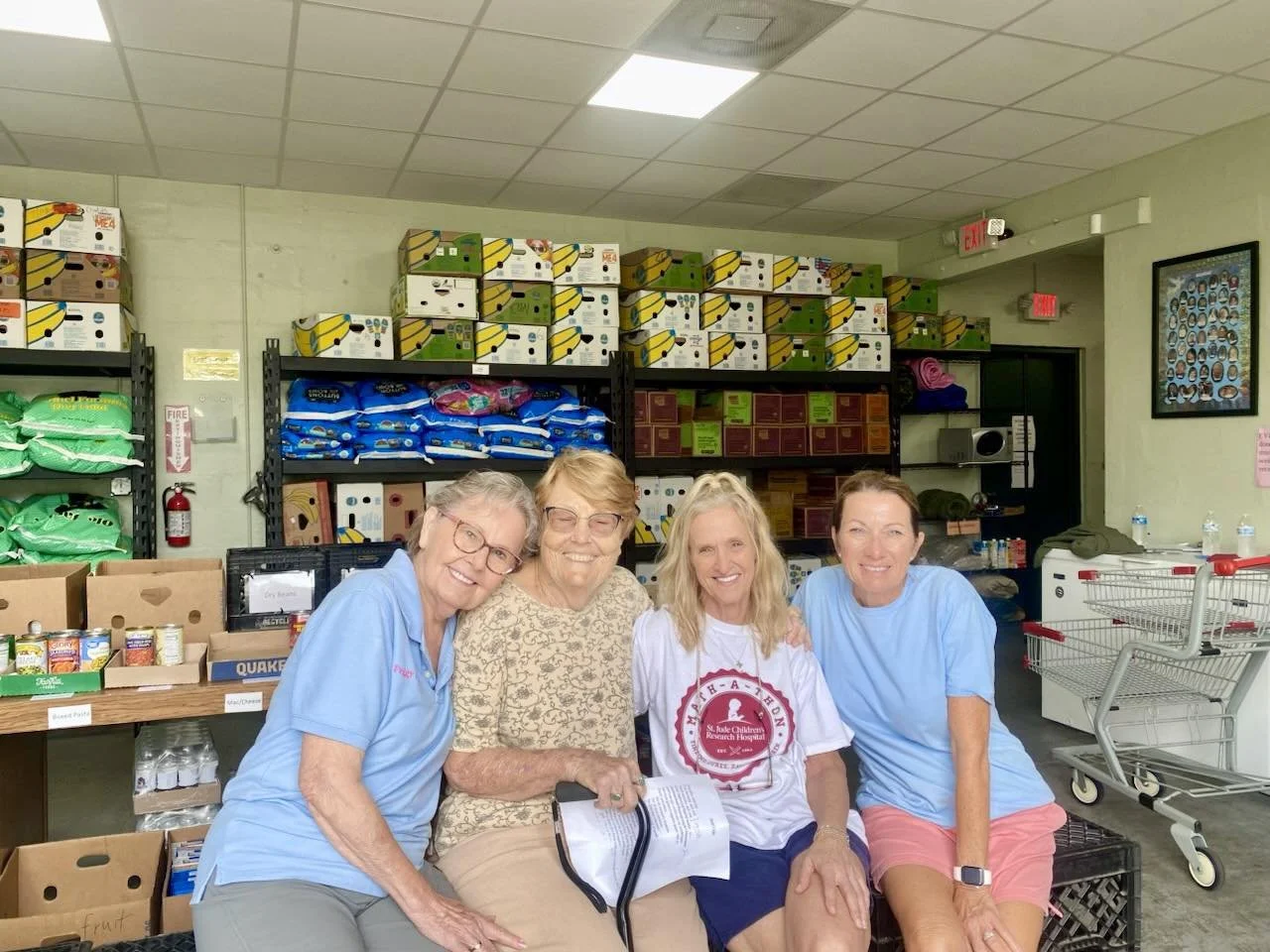 Four women sitting together in a food bank or storage room, smiling at the camera. Shelves behind them are stocked with boxes of food and supplies, and there is a shopping cart nearby.
