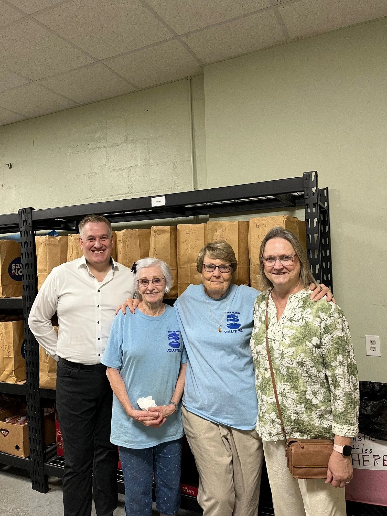 Four people smiling and posing for a photo inside a food pantry, with shelves of brown paper bags behind them. Two women are wearing light blue volunteer shirts and one woman has a brown purse.