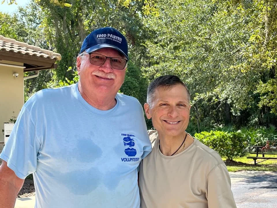 Two smiling men outdoors, one wearing a blue food pantry volunteer t-shirt and cap, and the other in a beige shirt, with trees and a park bench in the background.