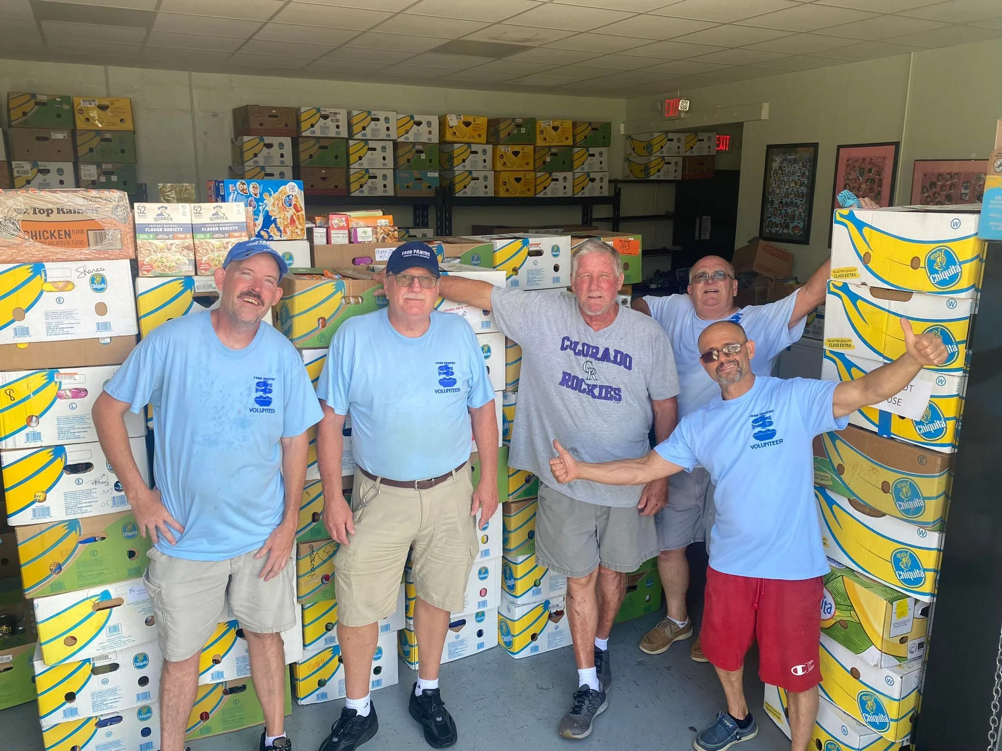 Five volunteers standing in front of boxes of bananas inside a food donation center or warehouse, smiling and giving thumbs-up.