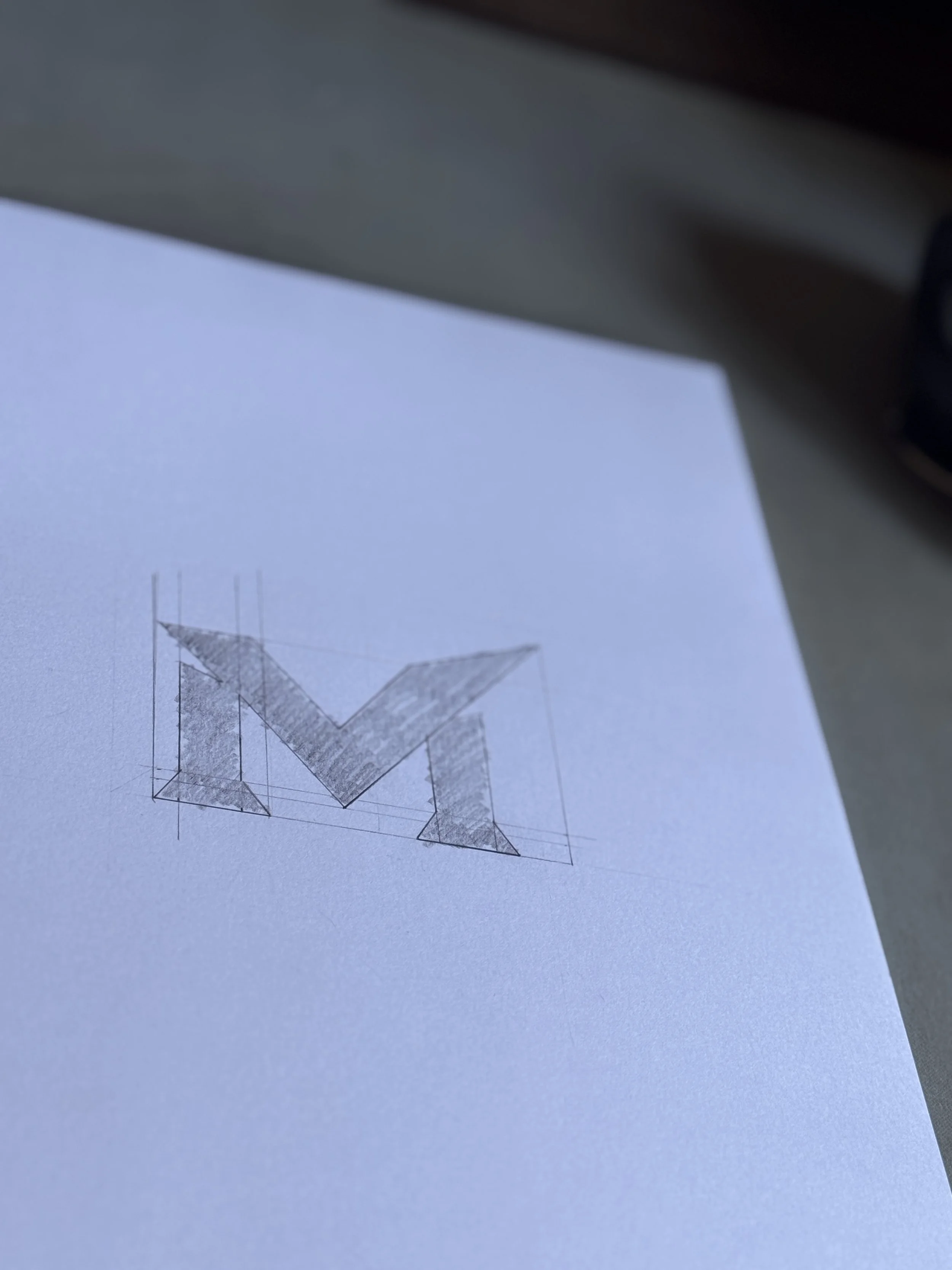 Sketch of a logo with a stylized letter 'M' on a piece of white paper.