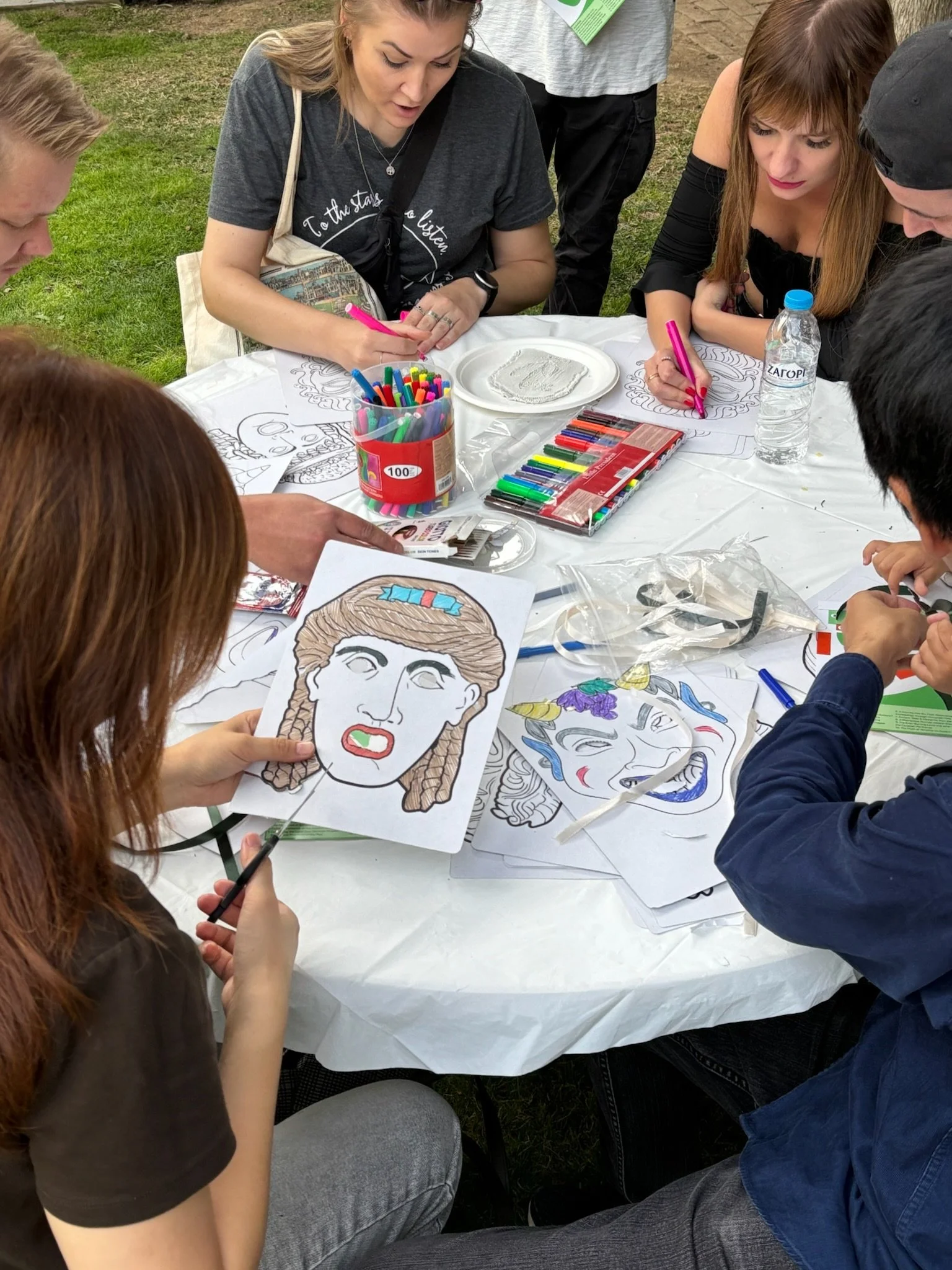 Group of people engaged in a face painting activity outdoors, with colorful markers, printed masks, and drawings on the table.