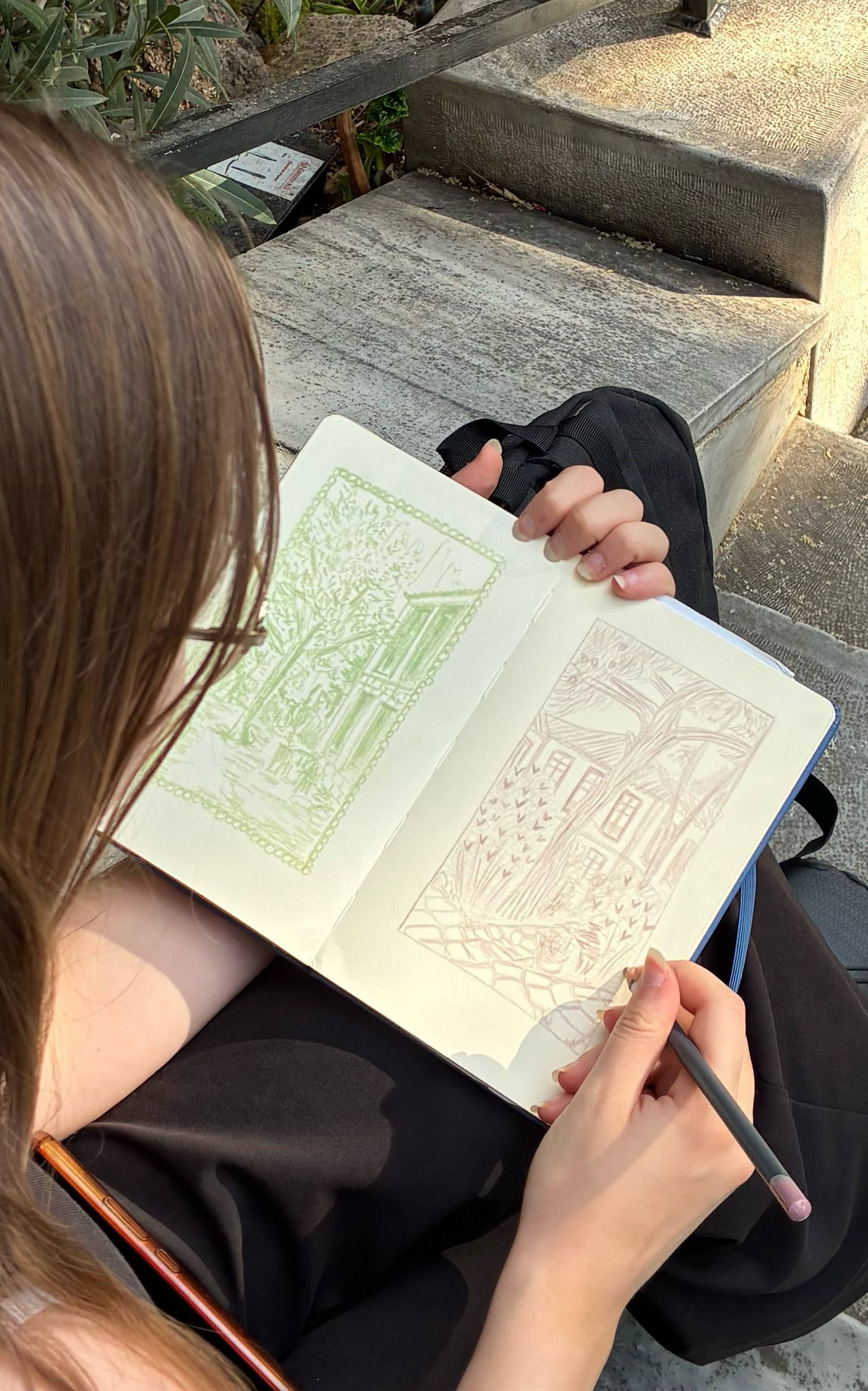 A person with brown hair and glasses sketching two landscape scenes in a sketchbook. One scene is in green ink, depicting trees and possibly a building with columns. The other scene is in reddish ink, showing a large tree, a building with multiple wi