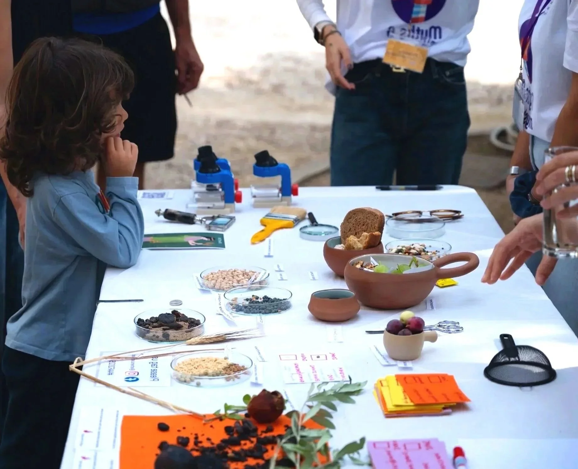 A young girl and several adults look at scientific specimens and tools on a table, including microscopes, petri dishes with various samples, and scientific notes, indicating a science exhibit or educational activity.