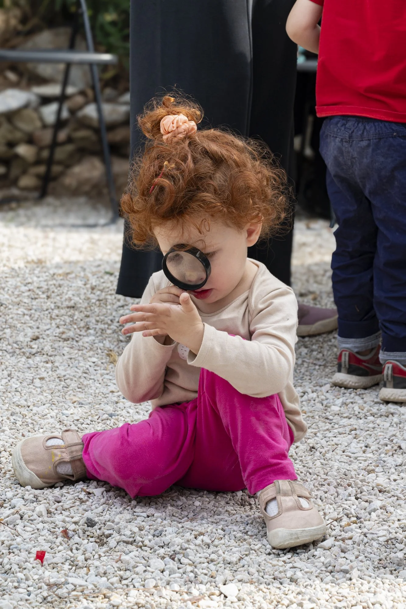 A young girl with curly red hair, wearing pink pants and beige shoes, sits on gravel while looking through a magnifying glass.