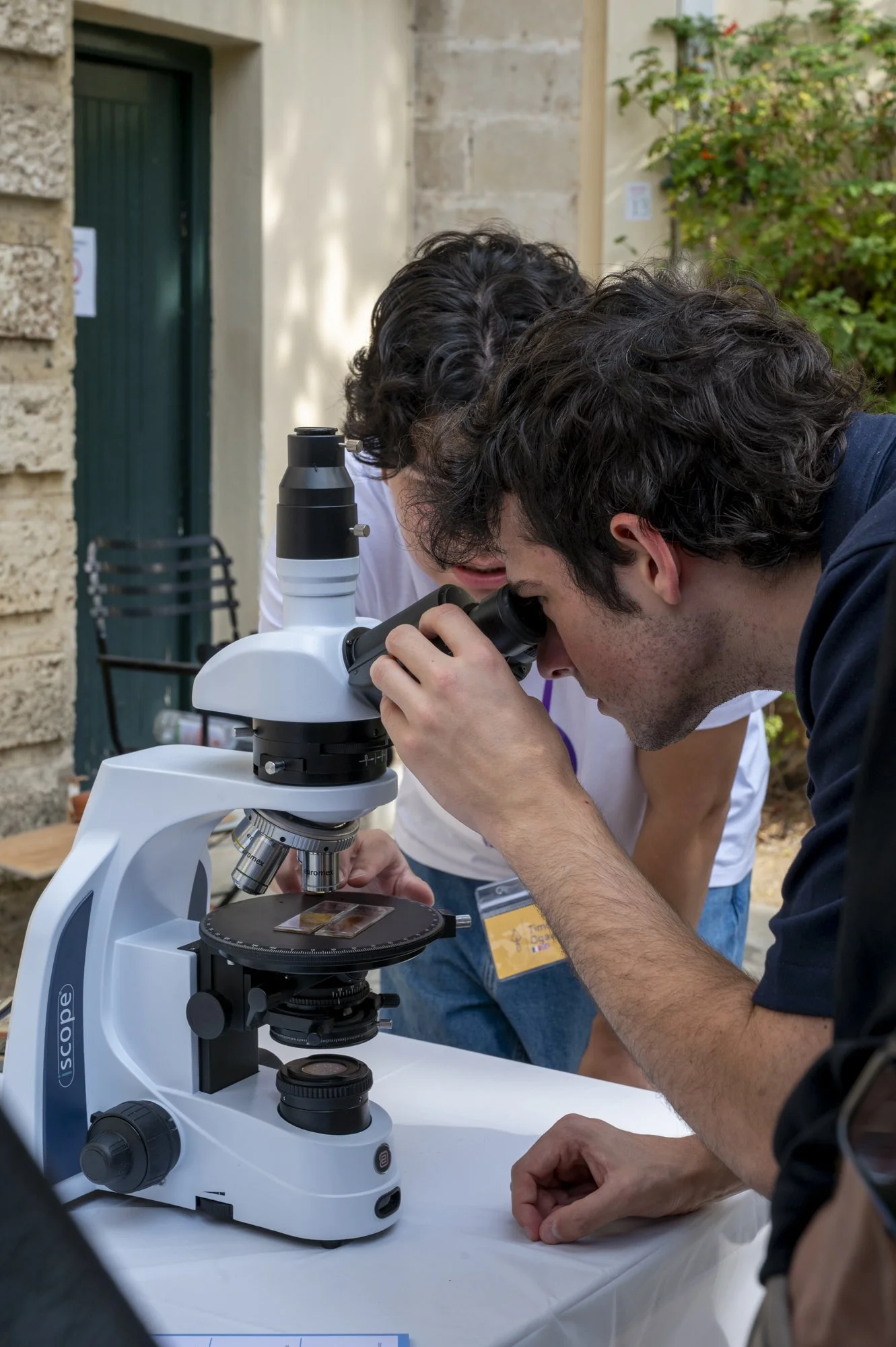 Two people examining a slide through a white and black microscope outdoors, with a beige brick wall and plants in the background.