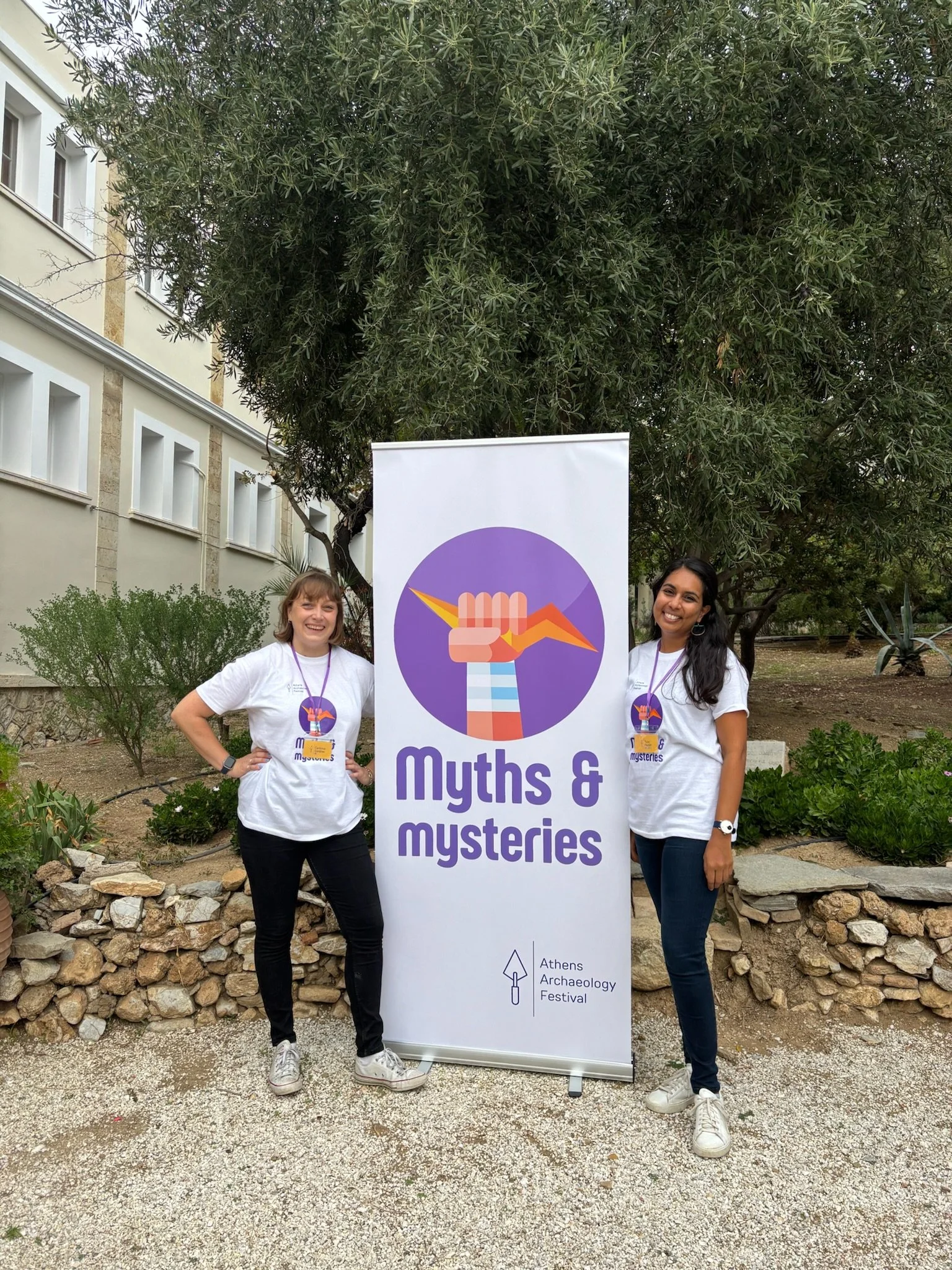 Two women standing outdoors next to a banner with the logo of Athens Archaeology Festival, which features a stylized hand holding an origami bird, with the event title 'Myths & Mysteries' and the festival name at the bottom. Both women are smiling and wearing white t-shirts and lanyards.