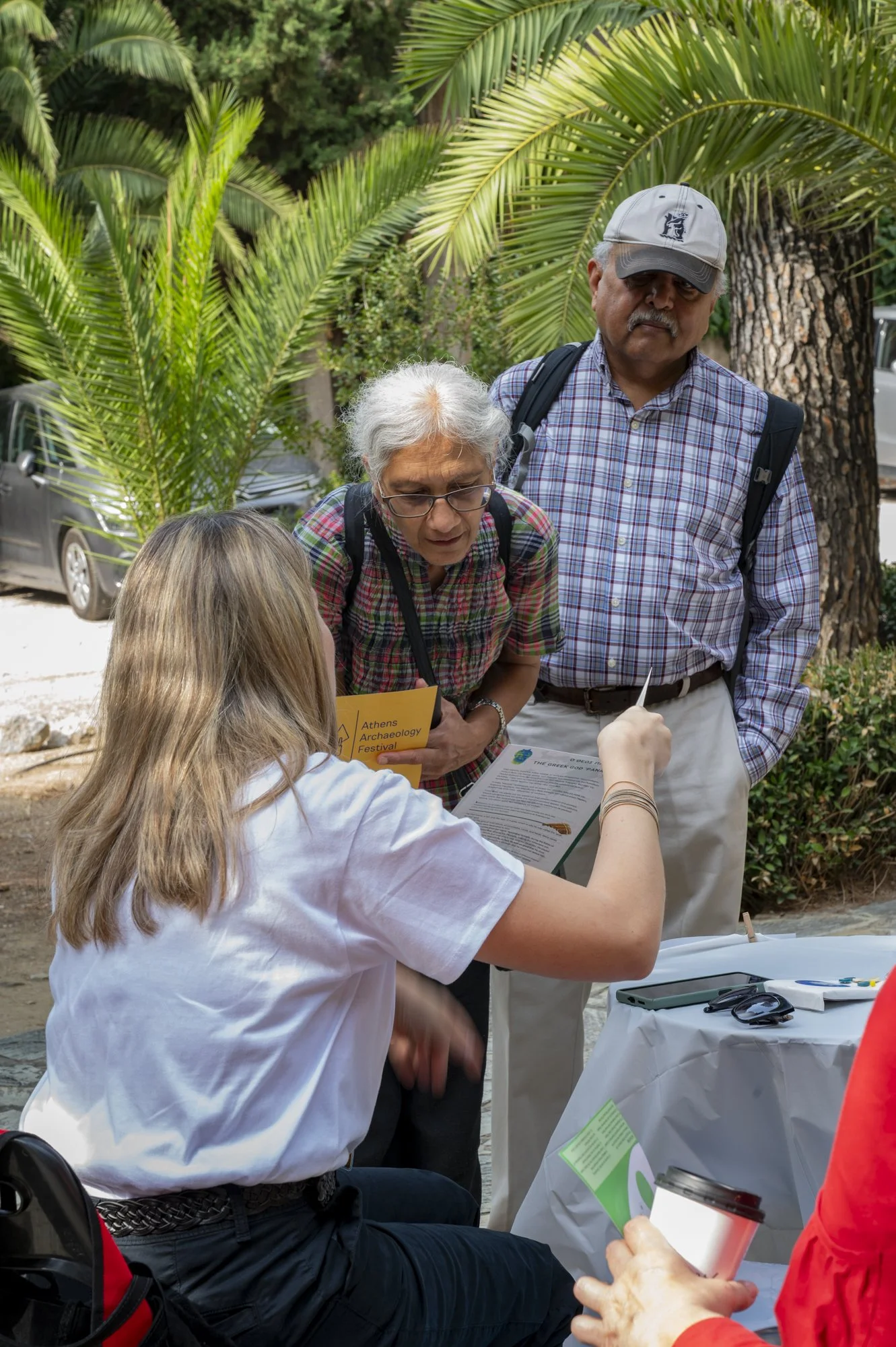 A group of three people talking at an outdoor event with green foliage in the background. One woman is seated, and two older adults, a woman and a man, are standing and engaging in conversation with her.