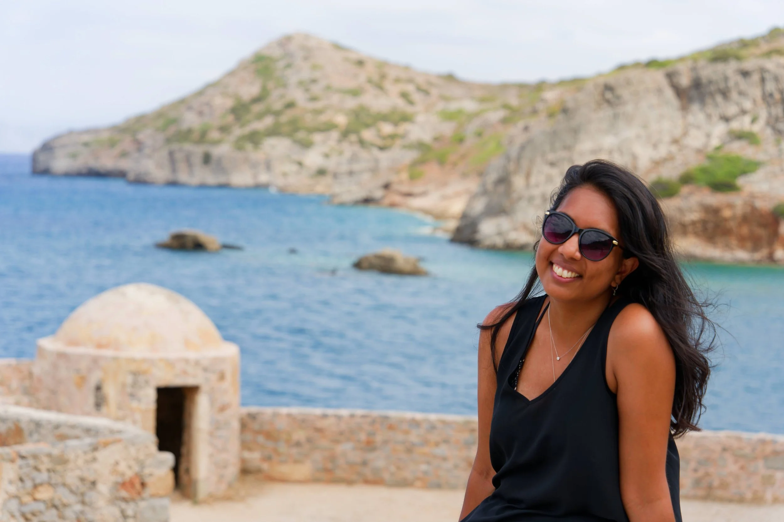 A woman with long dark hair wearing sunglasses and a black sleeveless top, smiling at the camera with a sea and rocky hills in the background.