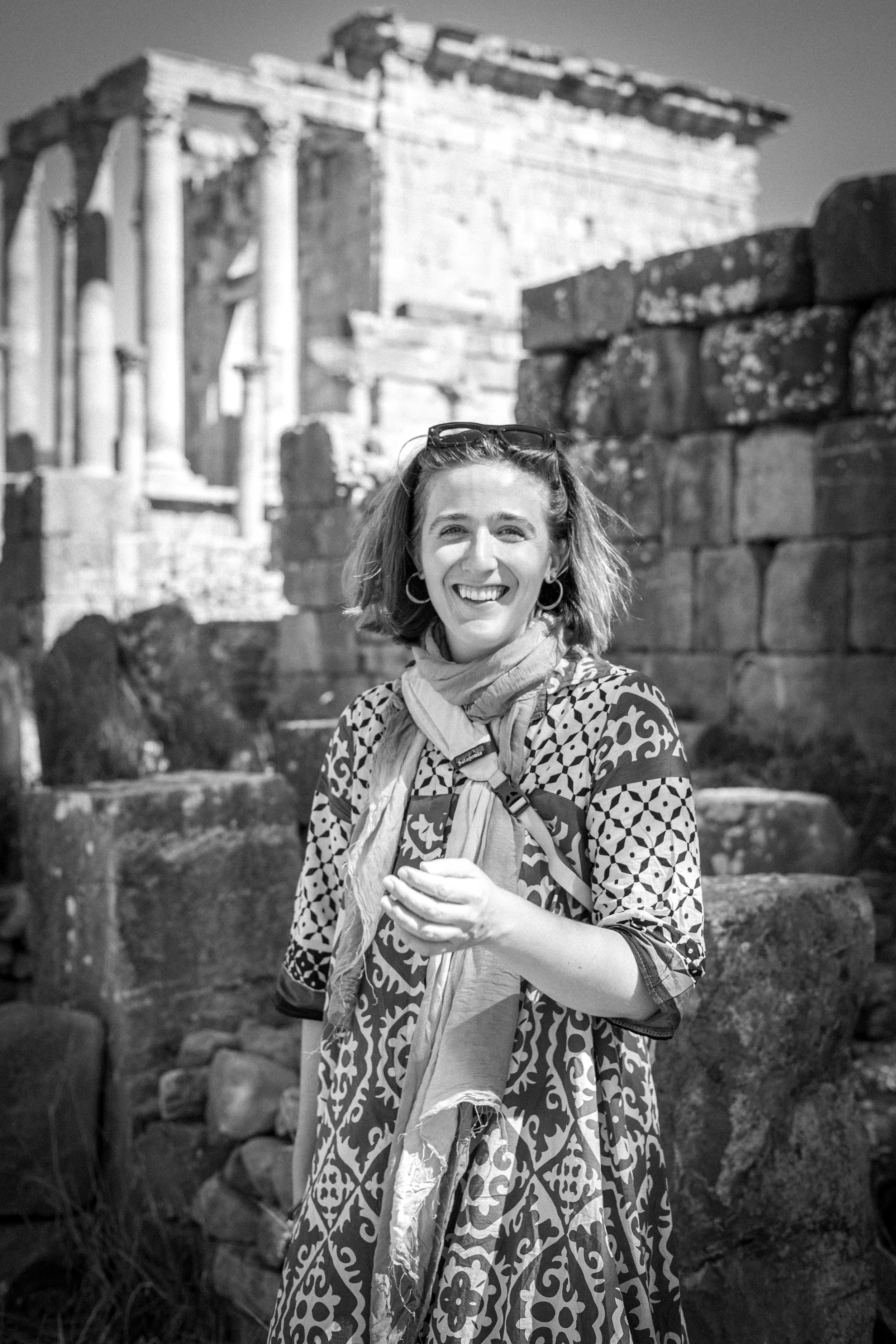 A woman with light hair, wearing earrings, a patterned dress, sunglasses on her head, and a scarf around her neck, smiling at the camera in front of ancient ruins made of stone pillars and blocks.