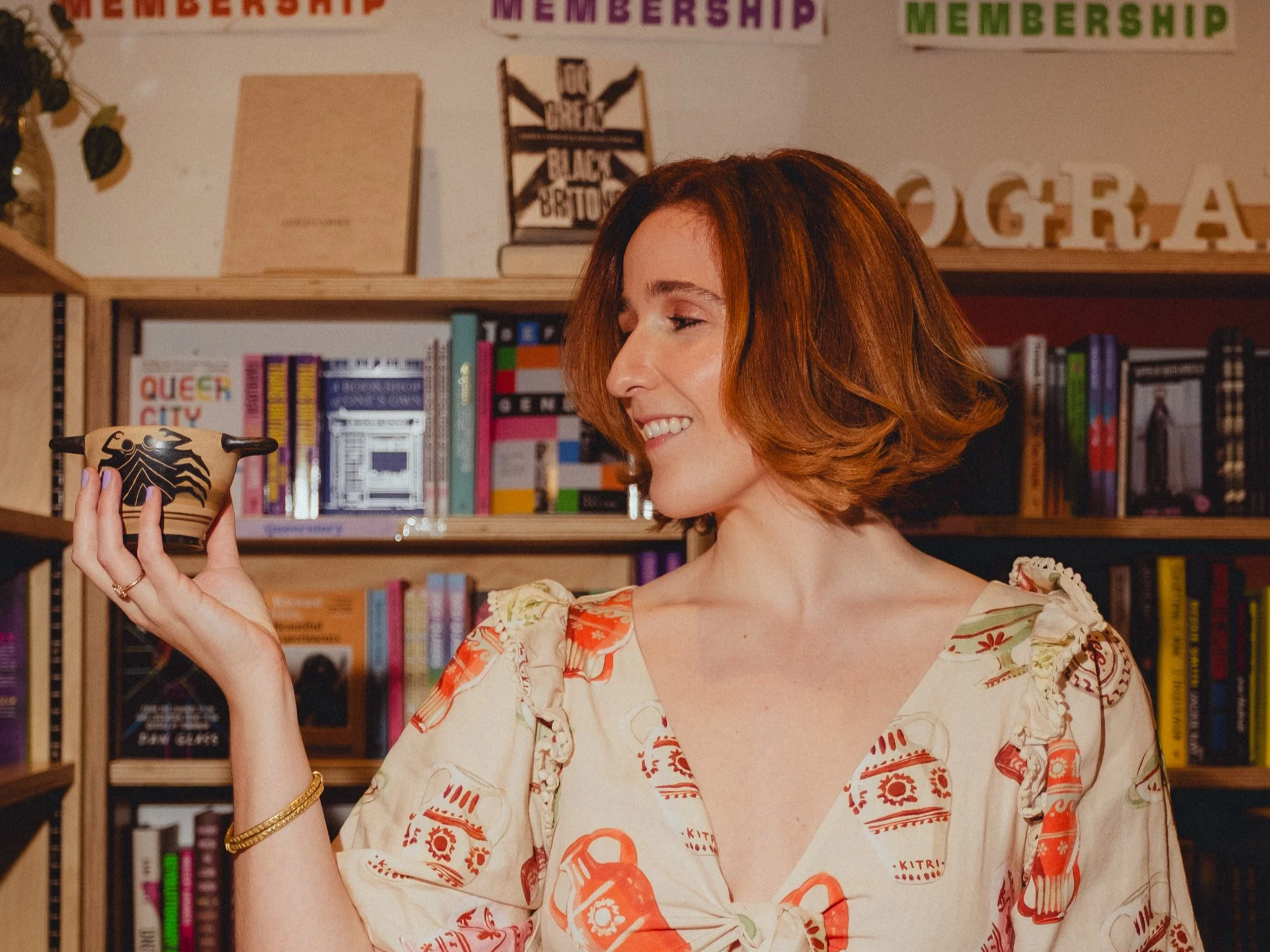 A woman with short red hair smiling while looking at a small decorative bowl she is holding in her hand. She is in a room with wooden shelves filled with books and colorful signs that read 'MEMBERSHIP' and 'GRA'. The woman is wearing a light-colored blouse with red and orange patterns.