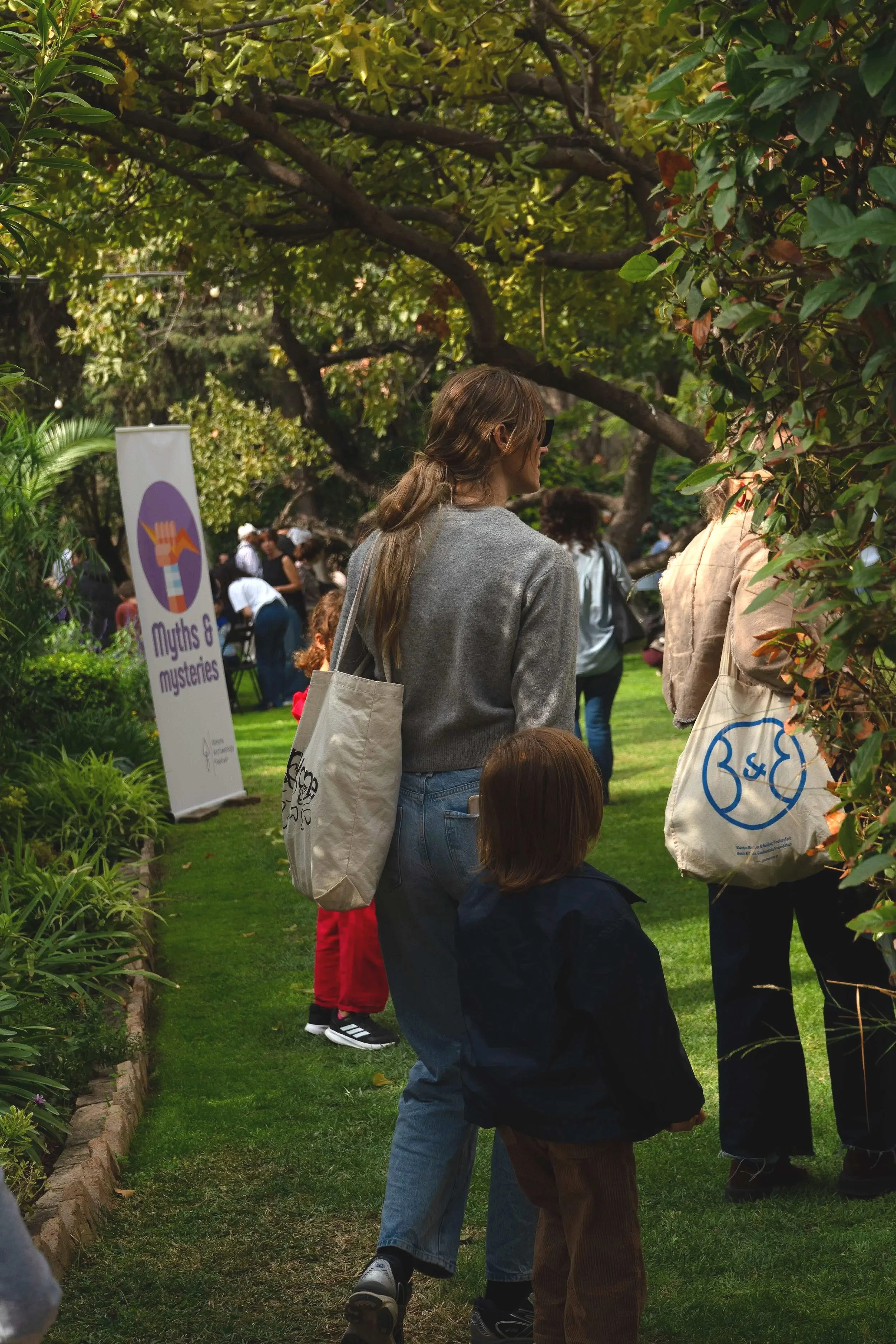 People at an outdoor event in a garden, including a woman with a tote bag and a young child, surrounded by trees and greenery, with a sign that says 'Myths & mysteries' in the background.