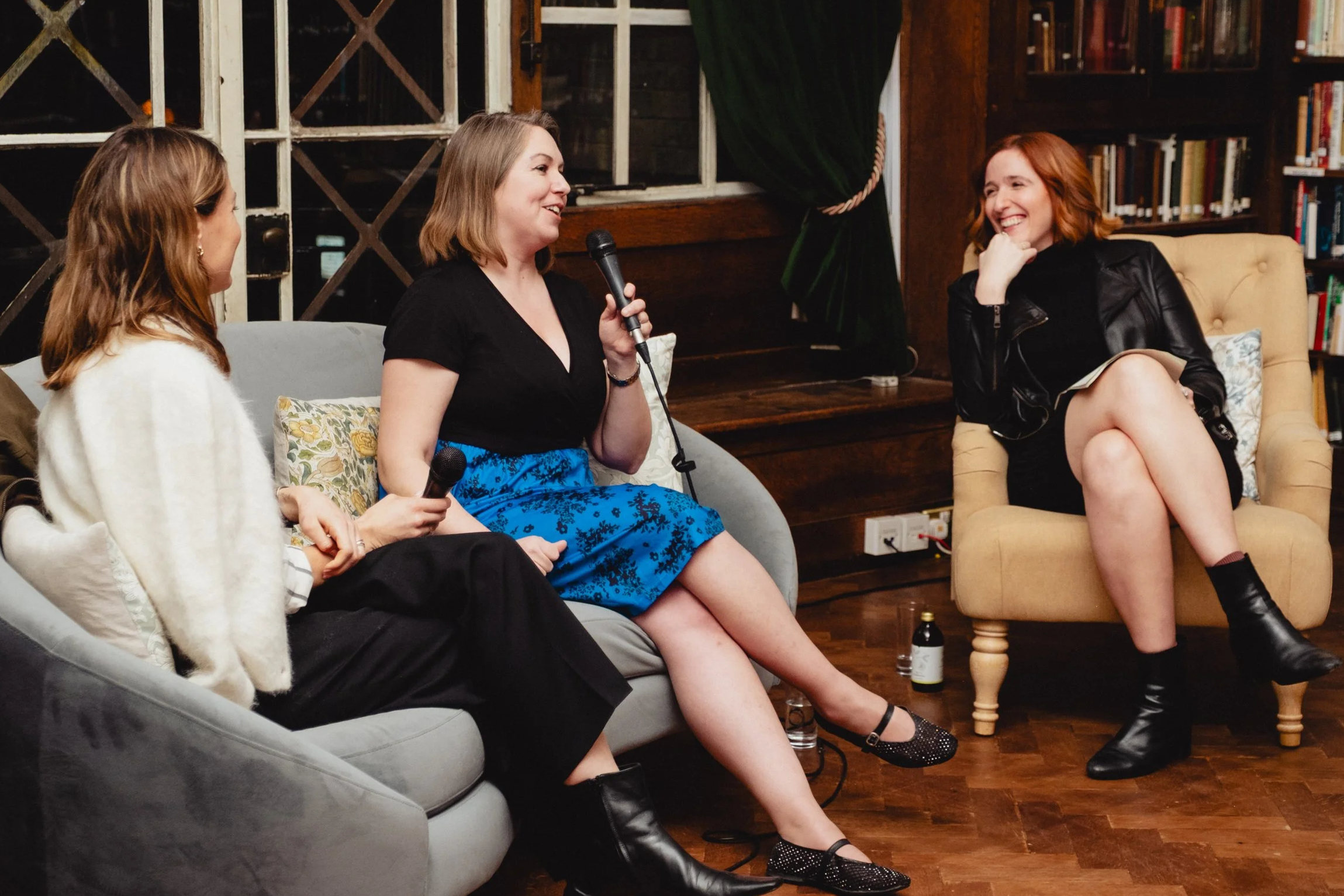 Three women sitting in a cozy library or bookstore, having a conversation during a casual event or interview. One woman is holding a microphone and speaking, while the other two women are listening and smiling.