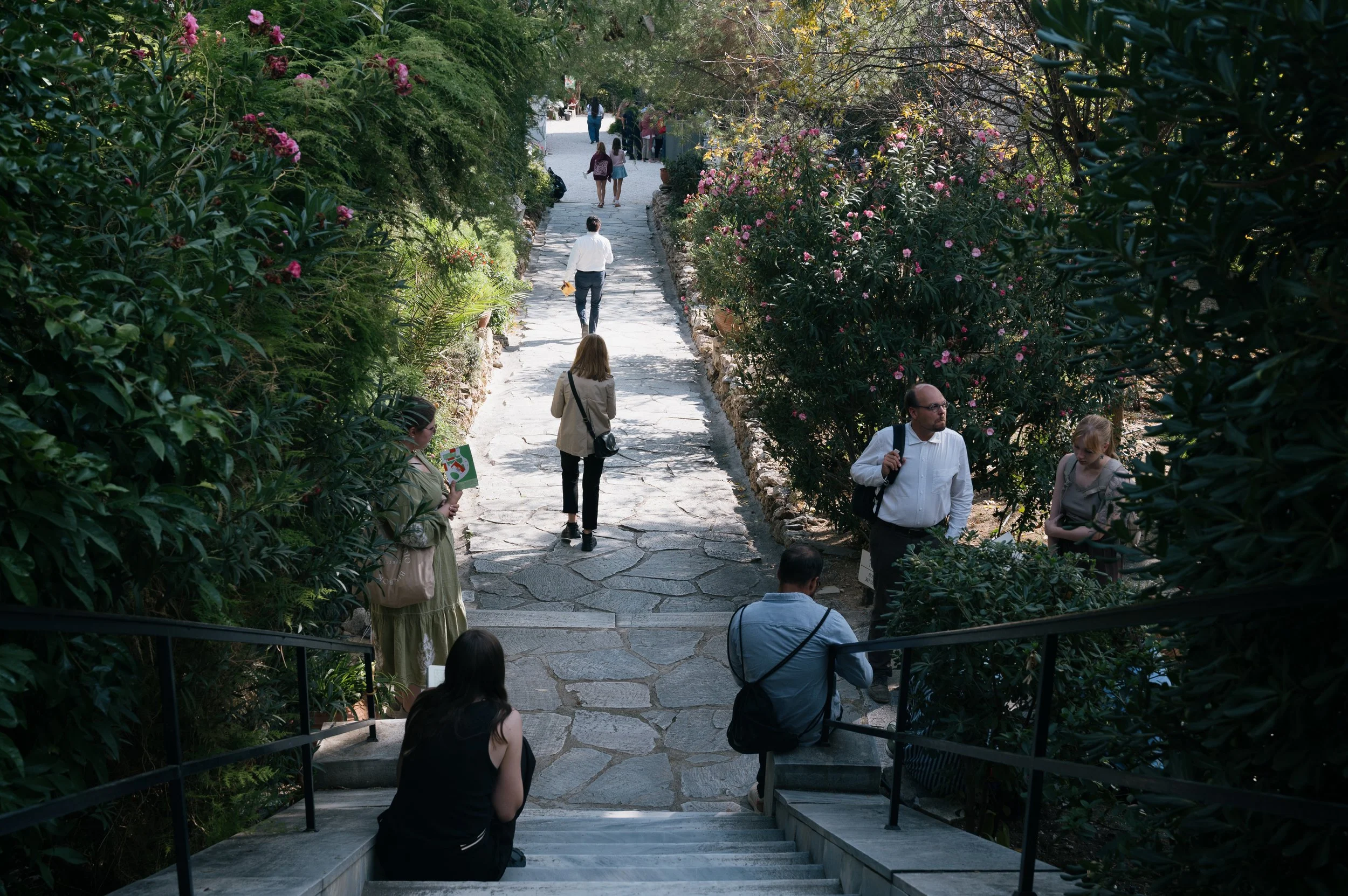 People sitting and walking on a stone pathway lined with lush green bushes and pink flowering plants, with some individuals carrying bags and books, in a garden or park setting.
