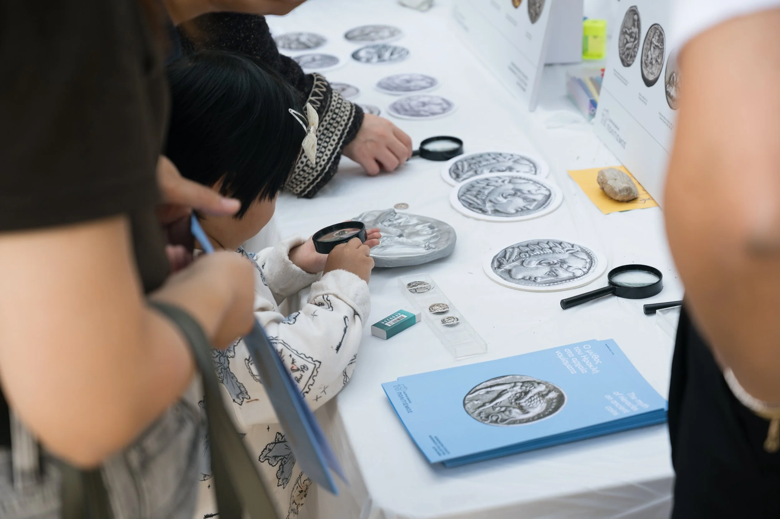 Children and adults examining and holding silver coins with magnifying glasses at a table displaying coin replicas, informational pamphlets, and a black test tube.