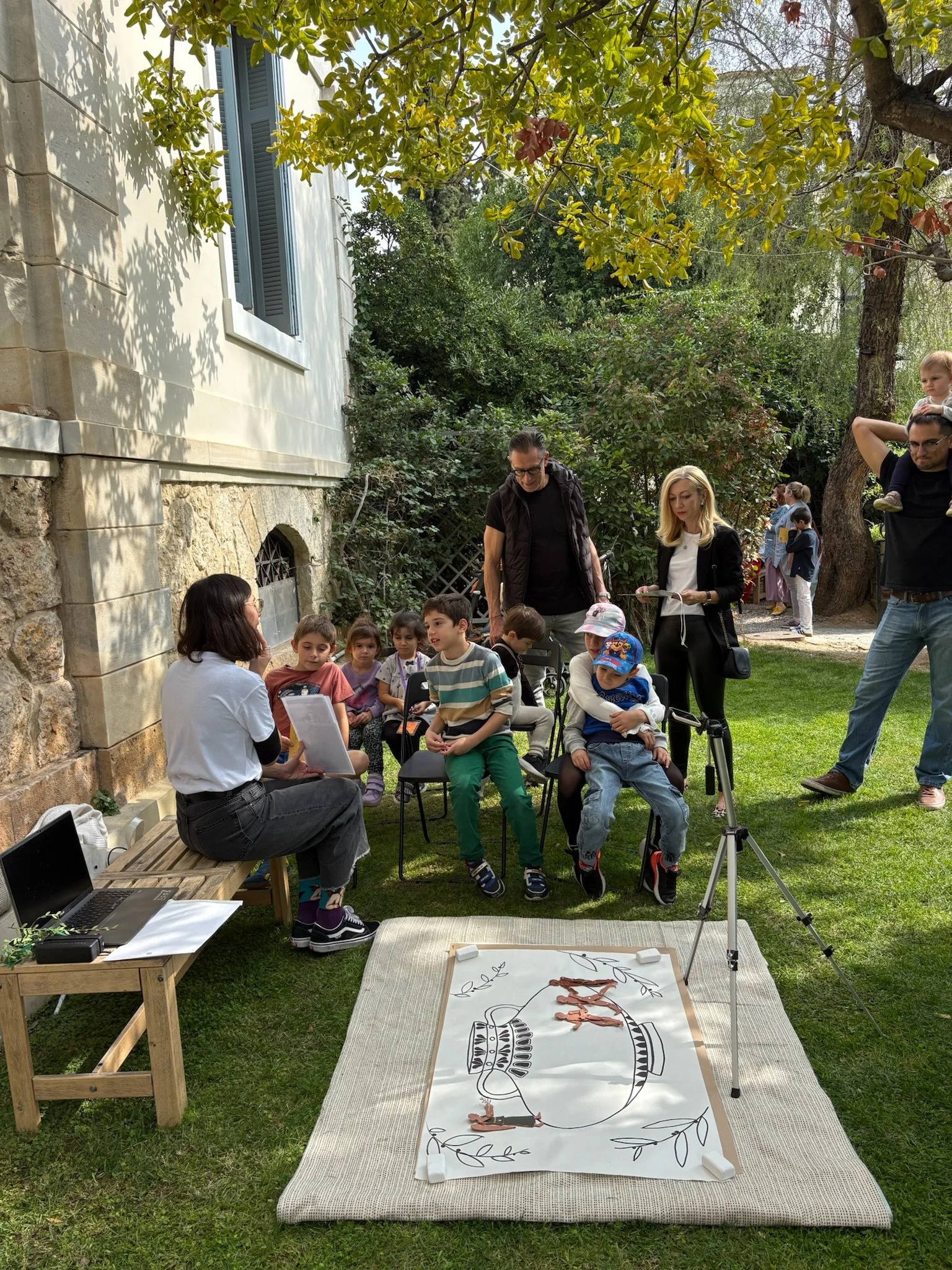 Group of children and adults gathered outdoors for an activity, with a large drawing of a fish and some goldfish figurines on a mat, and a camera on a tripod set up nearby.
