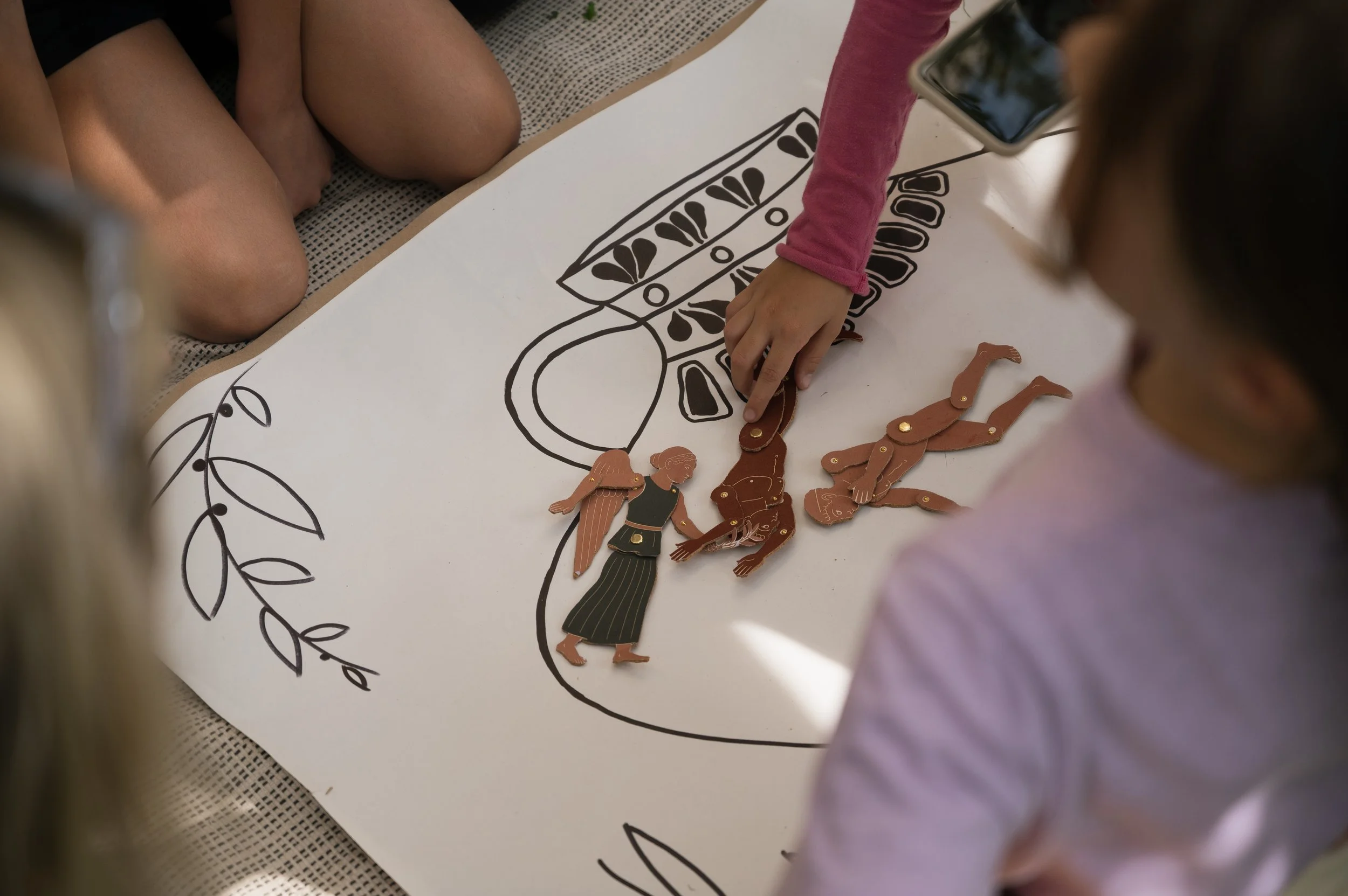 Children working on a collage art project with paper and cut-out illustrations of people, creating a scene on a large white sheet of paper.