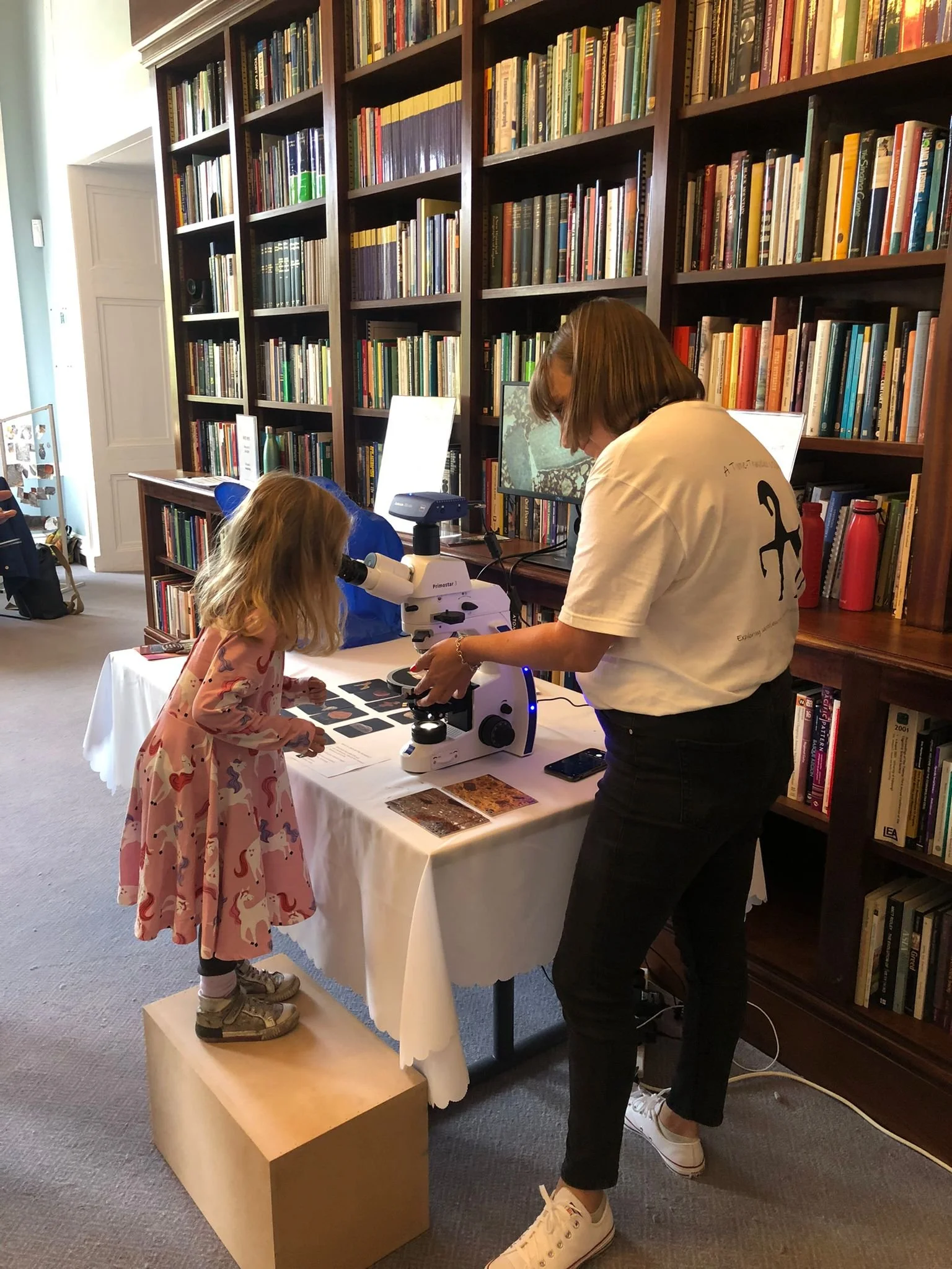 A woman and a young girl looking through a microscope at a table with educational materials in a library.