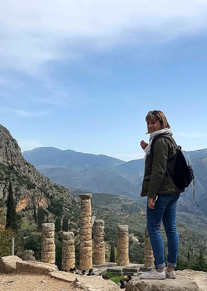 A woman with a backpack standing near ancient stone columns at an archaeological site, with mountains and a partly cloudy sky in the background.