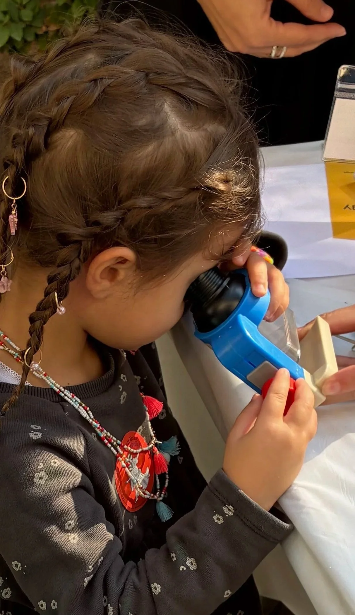 A young girl with braided hair, wearing a dark patterned shirt and colorful jewelry, looking through a blue telescope at a table with papers and a box.