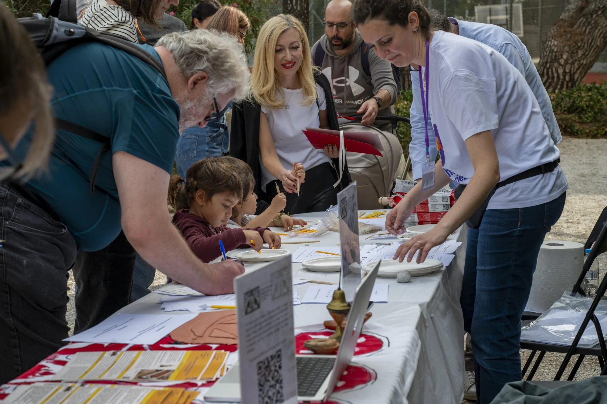 Group of people gathered around a table outdoors, engaging in activities like writing and shaping items, with children and adults participating, and informational signs and papers on the table.