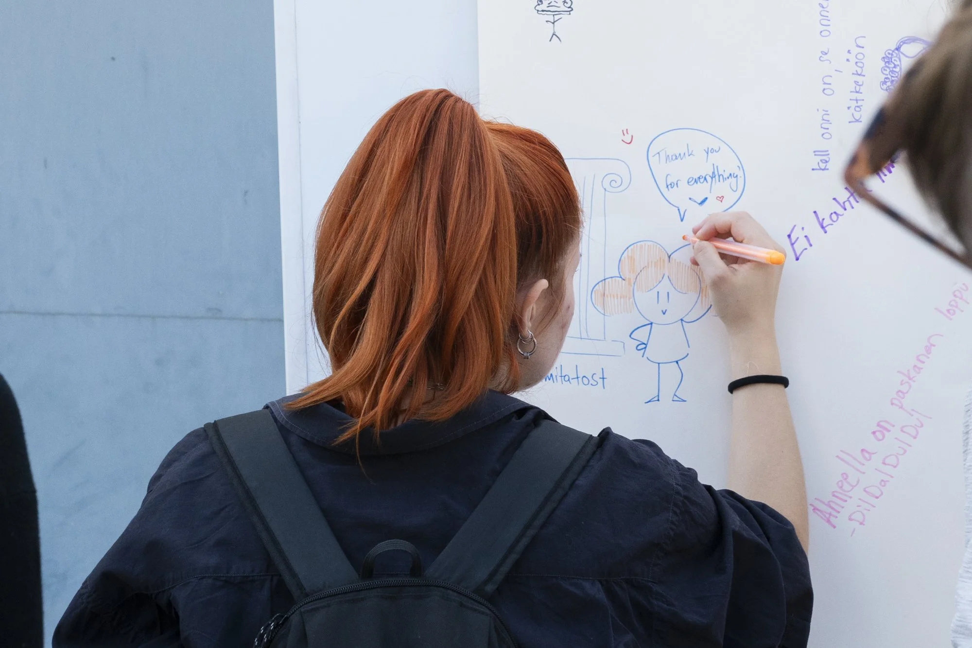 A person with red hair writing and drawing on a whiteboard, which has various doodles, messages, and notes in different colors, including a speech bubble and a cartoon character.