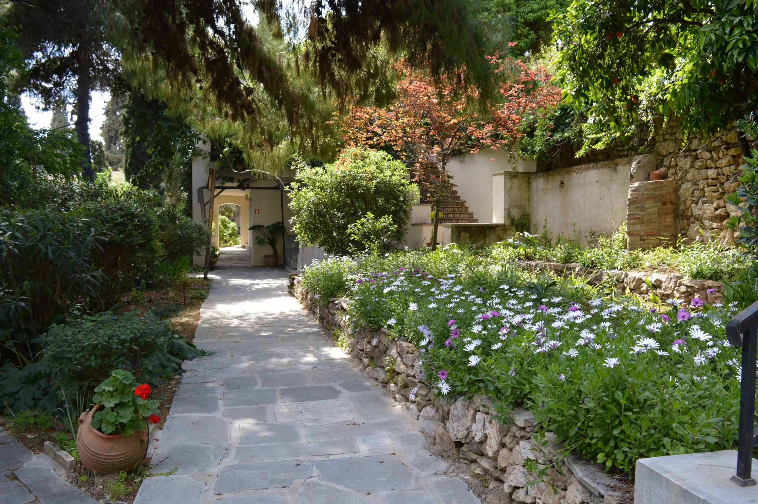 A stone pathway leading through a lush garden with colorful flowers, trees, and a white building with stairs in the background.