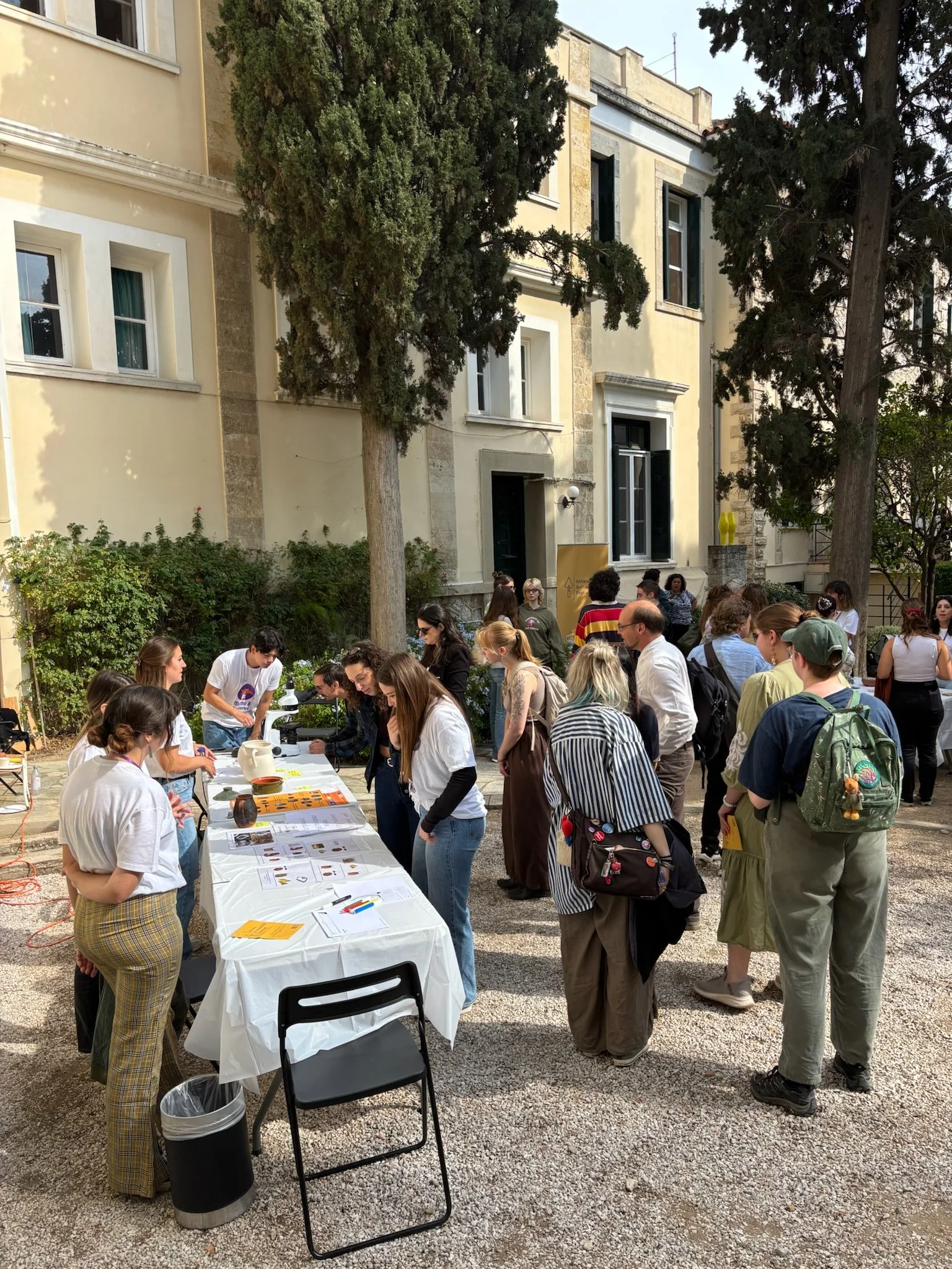 People gathered around tables with pamphlets and items at an outdoor event in a courtyard with trees and an old building in the background.