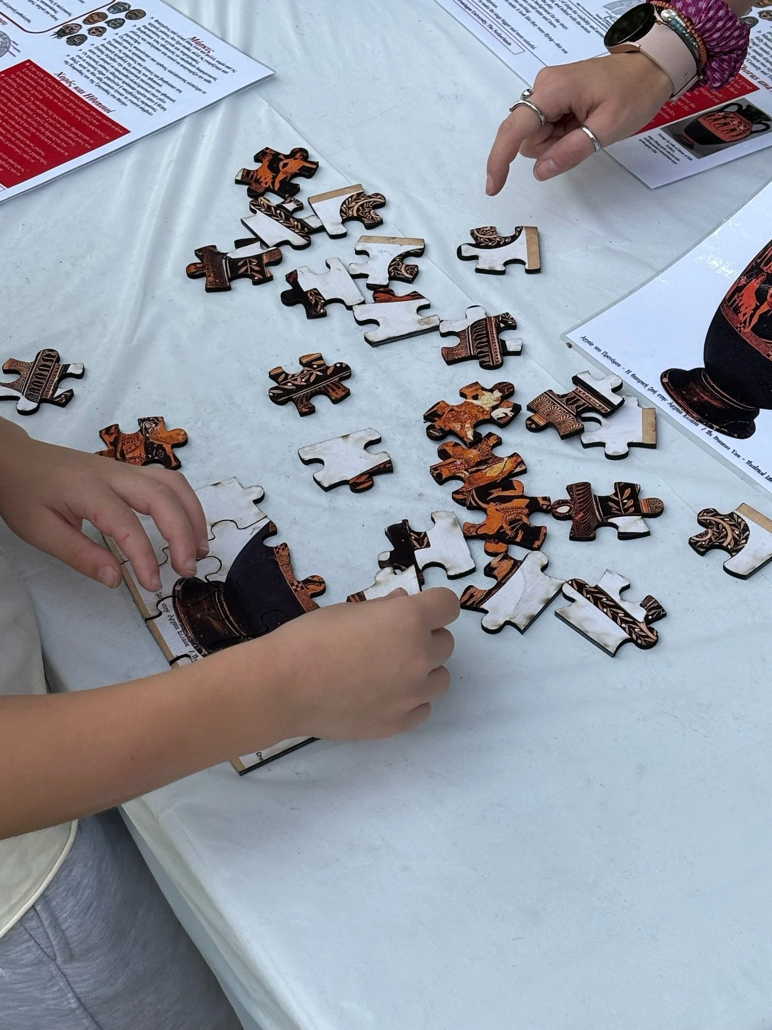 Two people assembling a wooden puzzle with 3D pieces on a white table, with printed informational pages nearby.