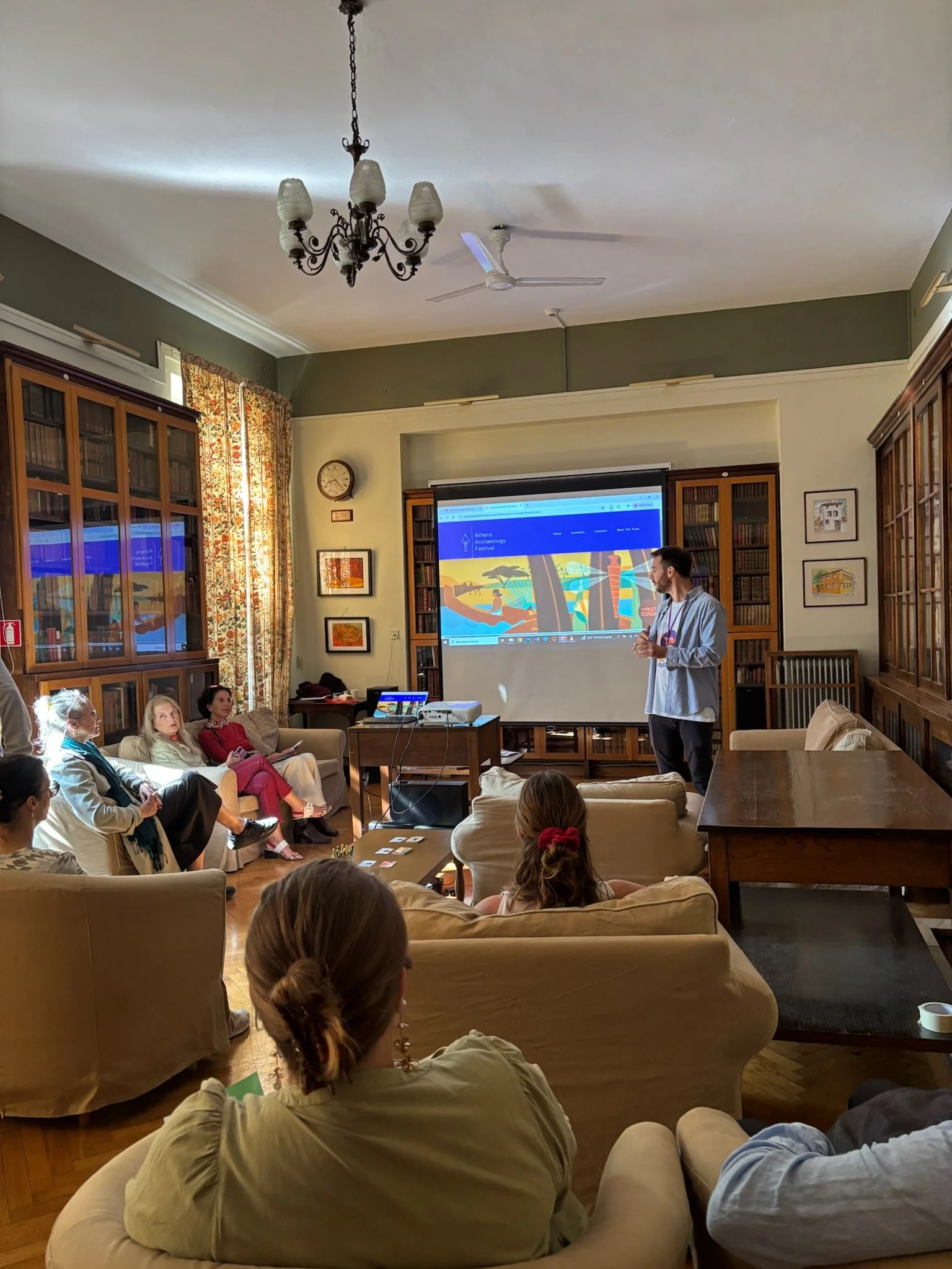 A man giving a presentation in a cozy room with wooden bookshelves, a projector, and an audience of women seated on sofas and armchairs.
