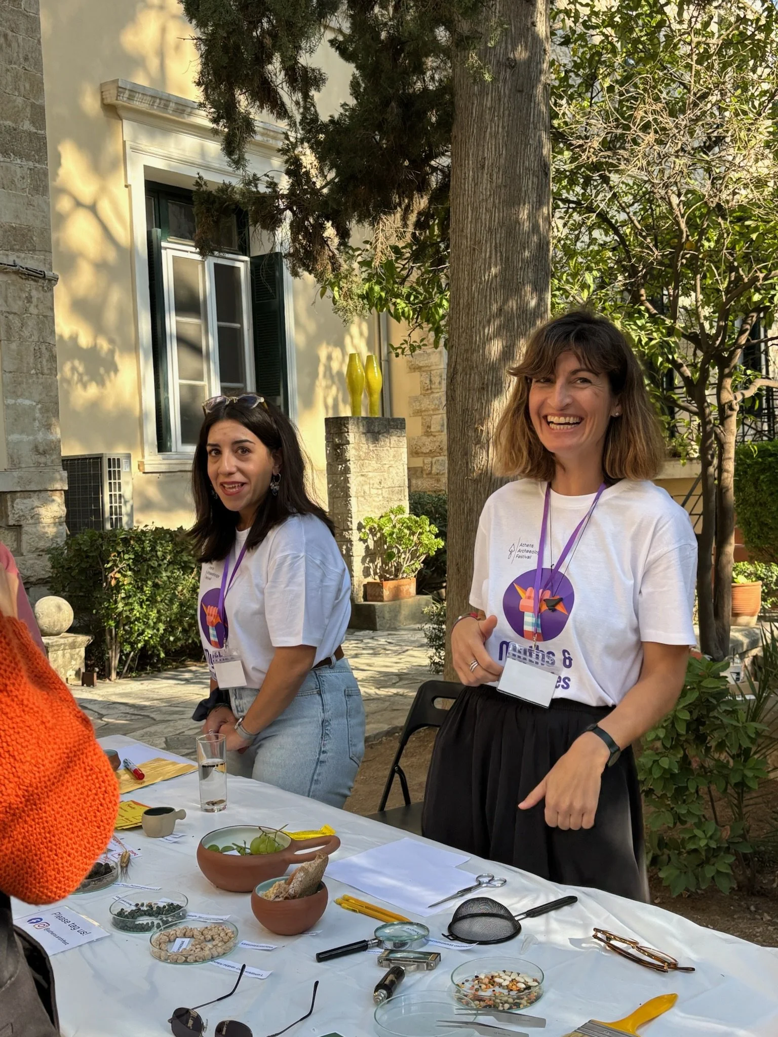 Two women standing behind a table outdoors, smiling. The table is decorated with various bowls and tools, suggesting a crafting or educational activity, with trees and a building in the background.