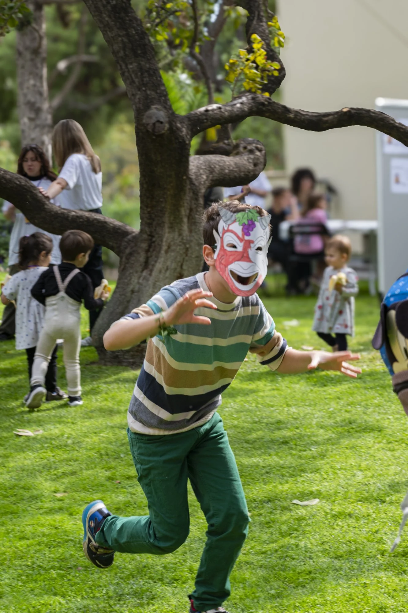 A boy wearing a laughing clown mask runs on a grassy yard surrounded by children and adults, with a large tree in the background.