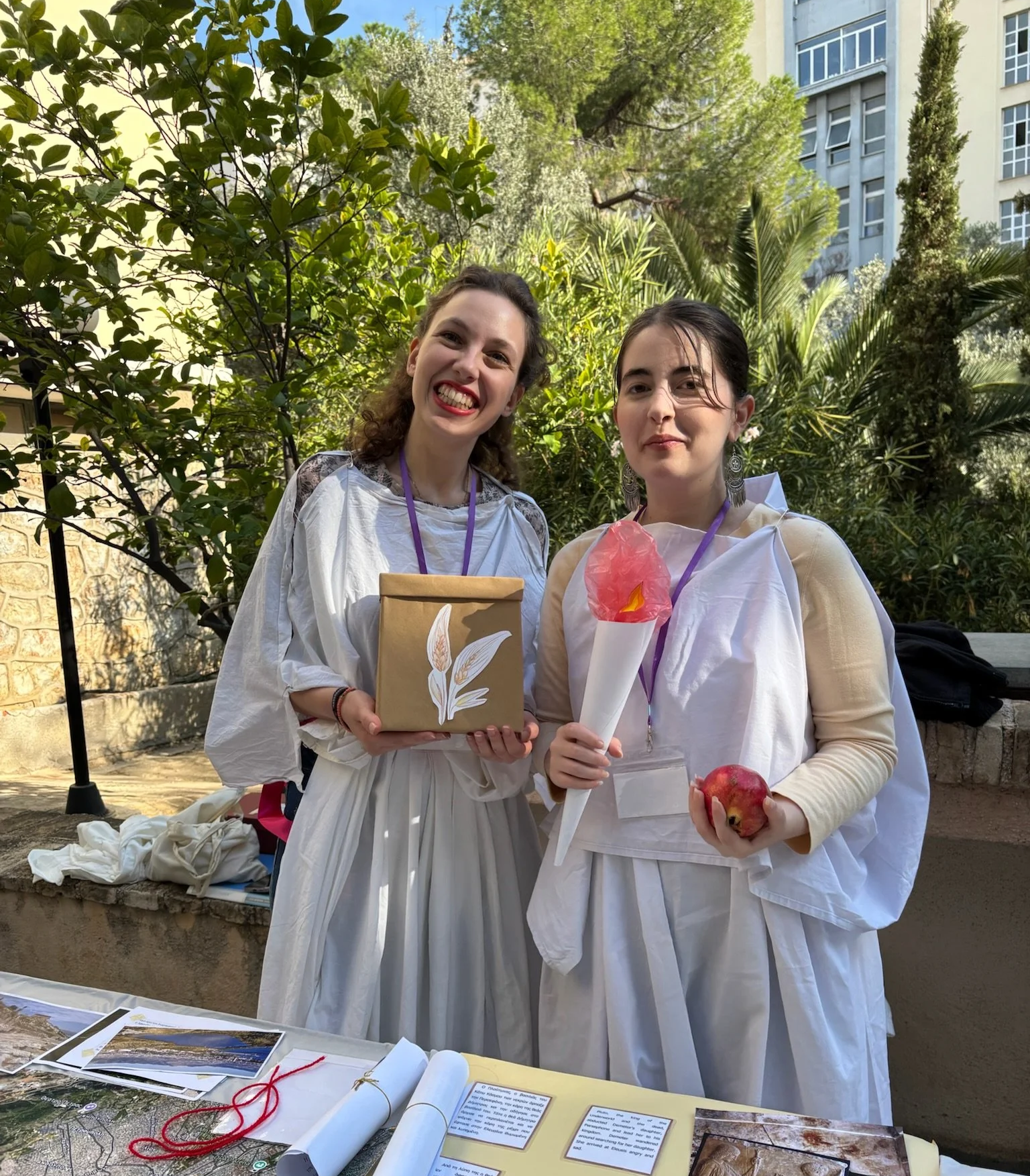 Two young women dressed in white stand outdoors, holding gifts and props, smiling. One holds a wrapped gift with a leaf design, the other holds a torch-shaped object and an apple. They are surrounded by green trees and plants, with a building in the background.