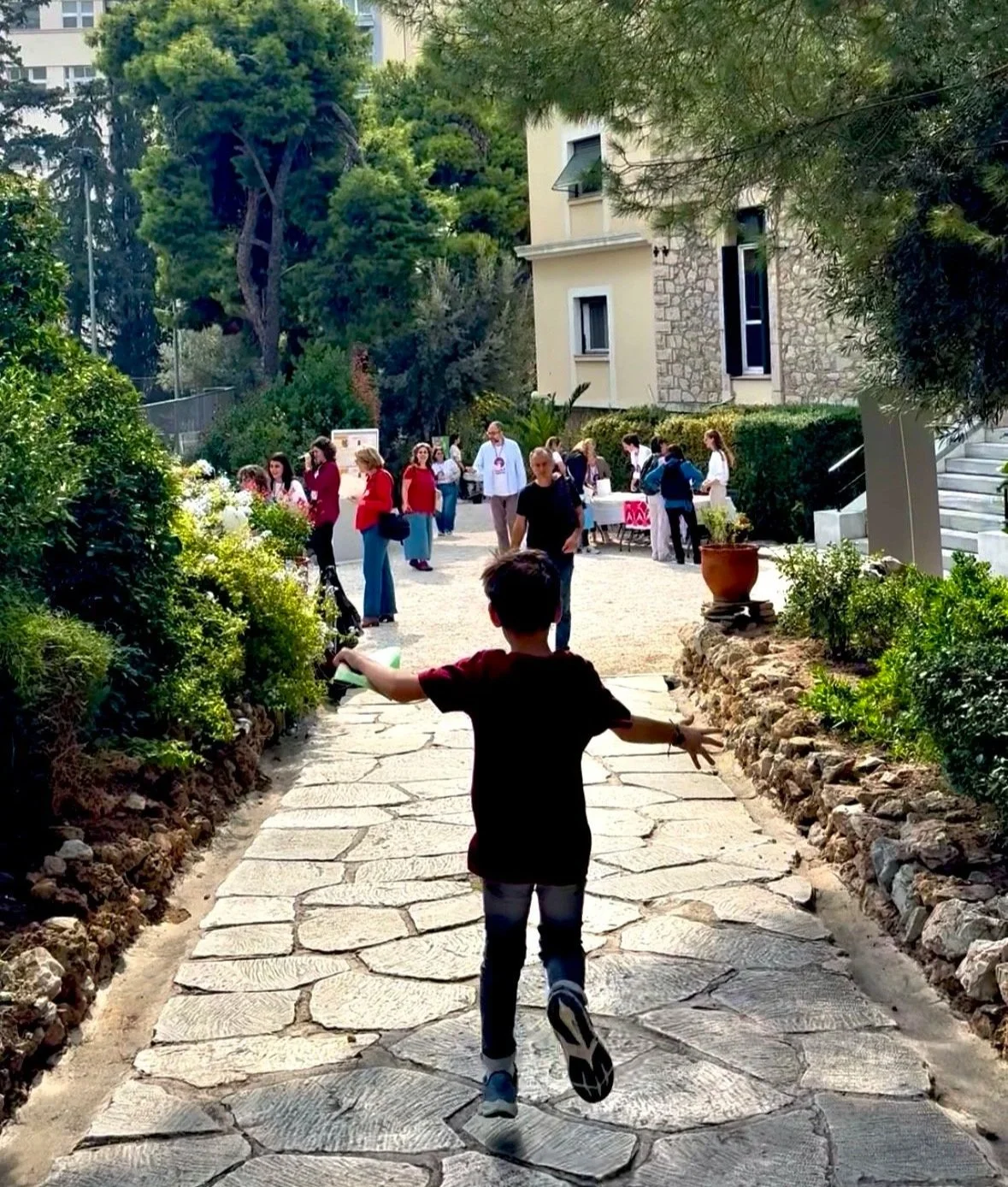 A young boy running toward a group of people gathered outdoors on a stone pathway, with plants and trees surrounding the area and a residential building in the background.