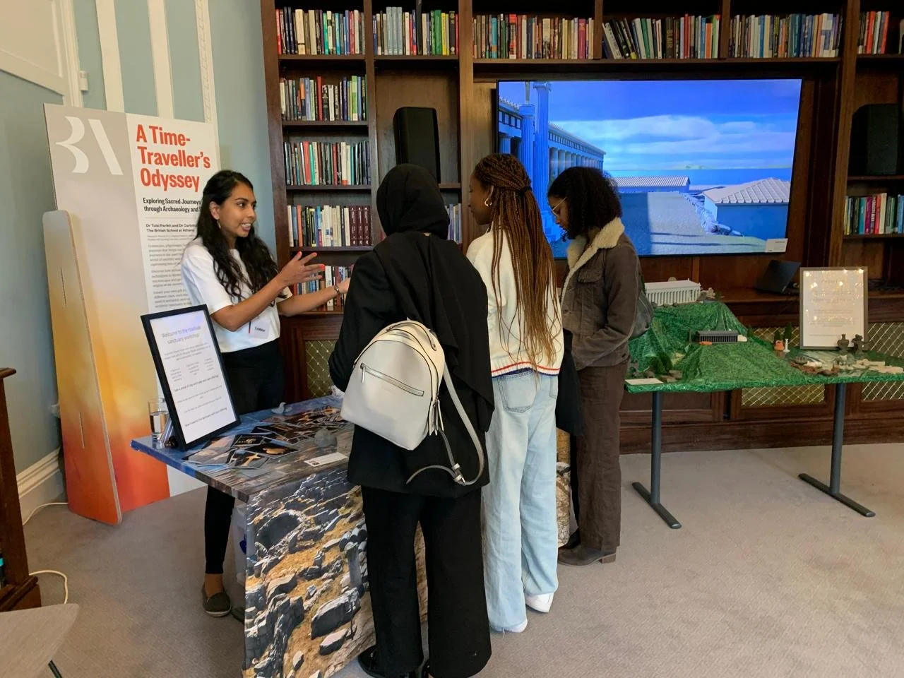 A group of four women at a booth in a bookstore or library, engaging with a young woman who is explaining something. The booth has a large sign titled 'A Time-Traveller's Odyssey,' and there are books and a display on a tabie to the right with a green fabric backdrop. A large screen showing a digital scene of a bridge and buildings is mounted behind them, and bookshelves filled with books are on the wall.
