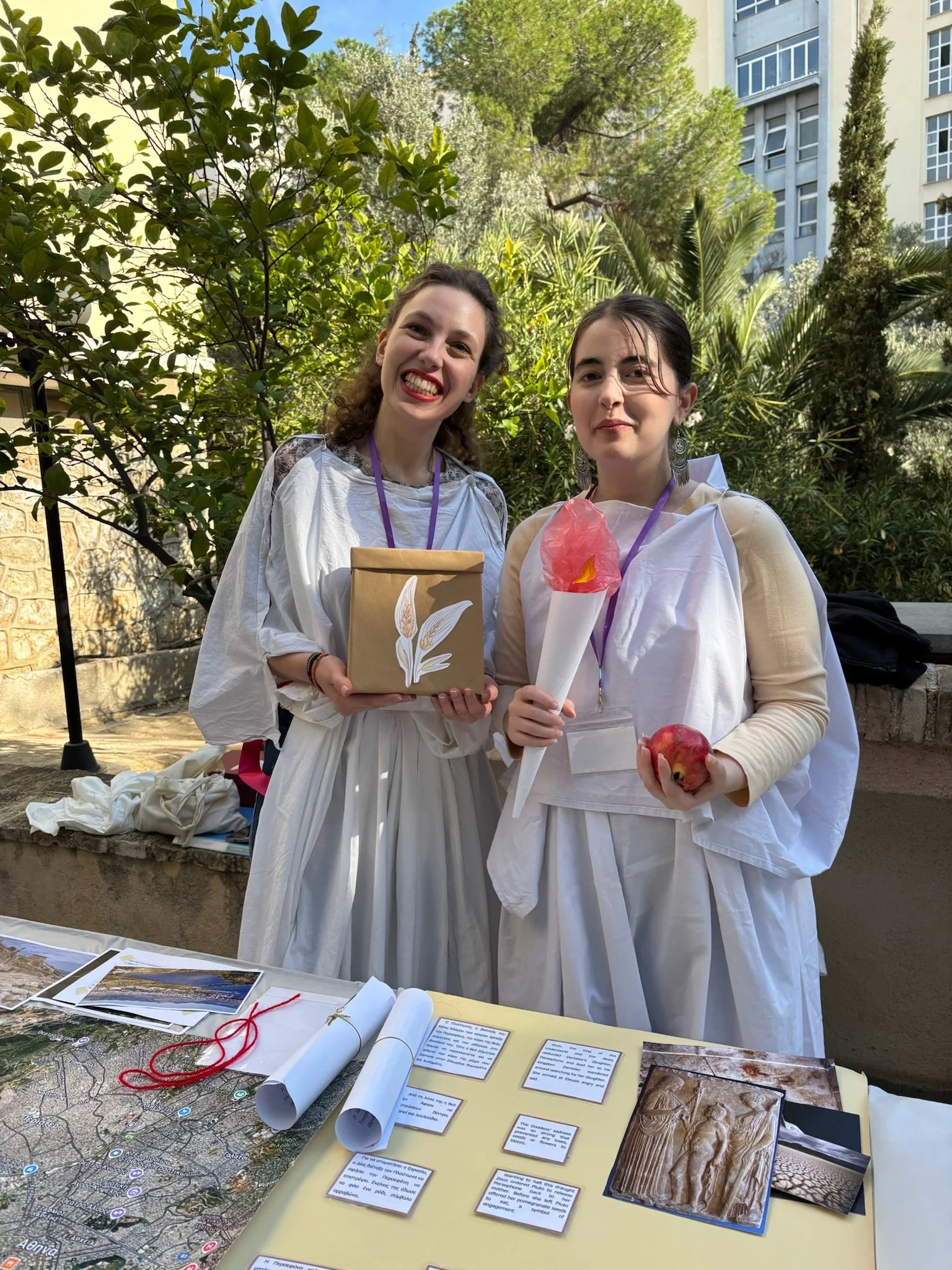 Two women dressed in white robes stand outdoors surrounded by green trees and plants. They are smiling and holding objects; one holds a small wrapped gift, and the other holds a decorative torch and an apple. A table in front of them displays papers,