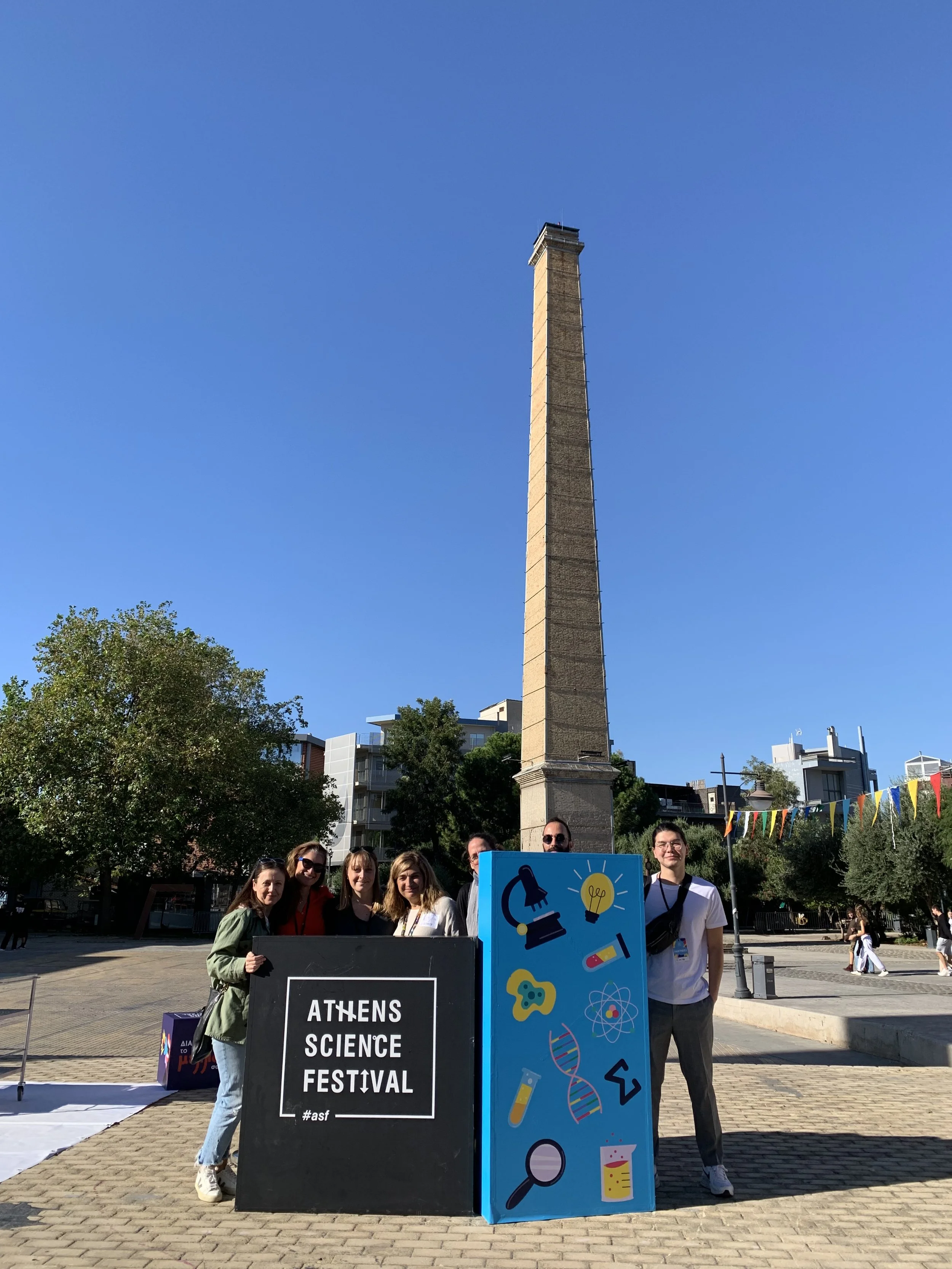 Group of people standing behind signs at the Athens Science Festival, with the ancient Athens Tower in the background on a sunny day.