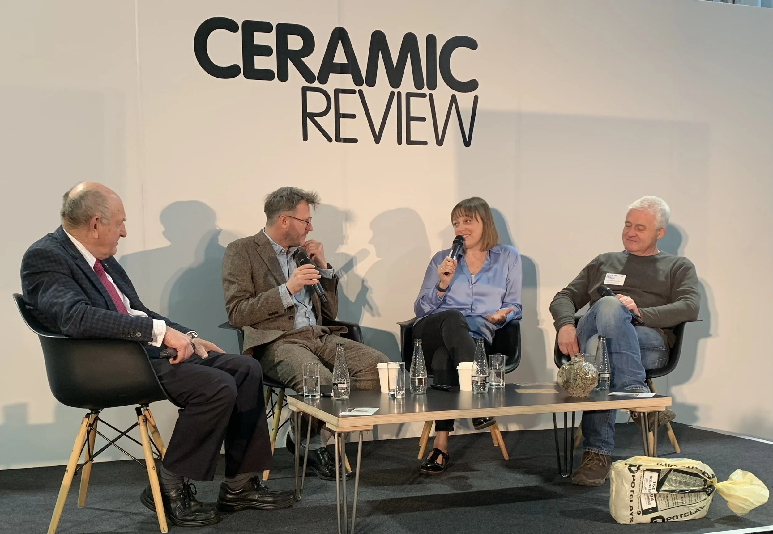 A panel discussion at a ceramic review event with four people seated on stage, each holding a microphone. There are tables with bottled water and paper cups, and a bag on the floor. The background has a large sign reading 'CERAMIC REVIEW'.