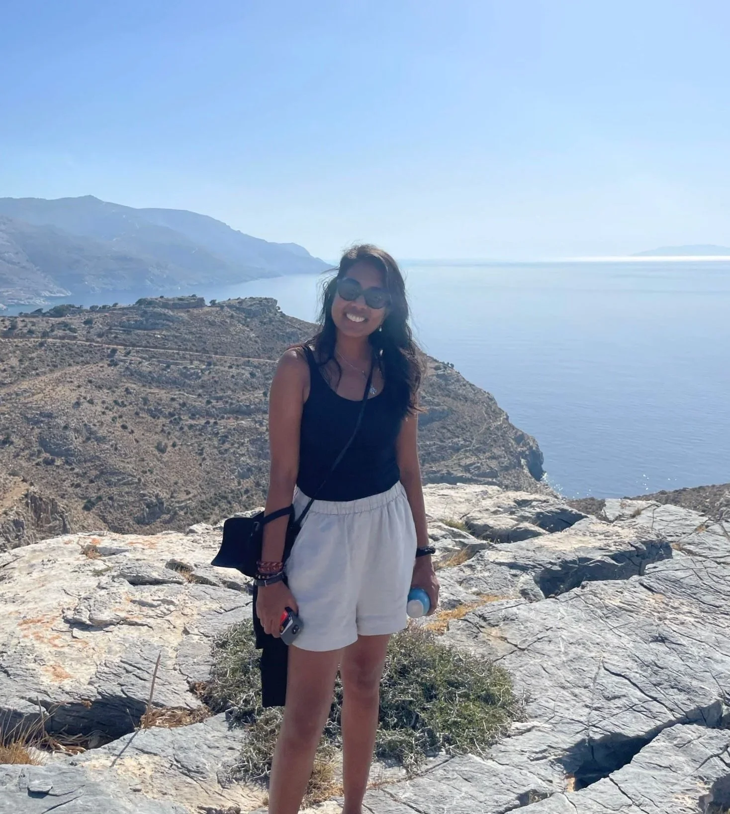 A woman standing on rocky terrain near the coast, smiling, wearing sunglasses, a black tank top, and white shorts, with a scenic view of mountains and water in the background.