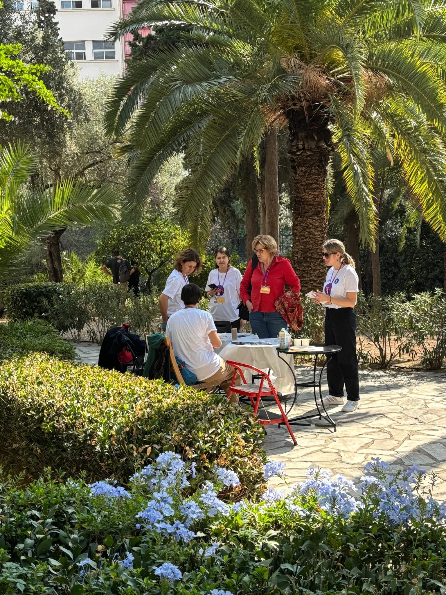 Group of five people gathered around a table in a lush garden with large palm trees and flowering bushes, engaged in a discussion or activity.