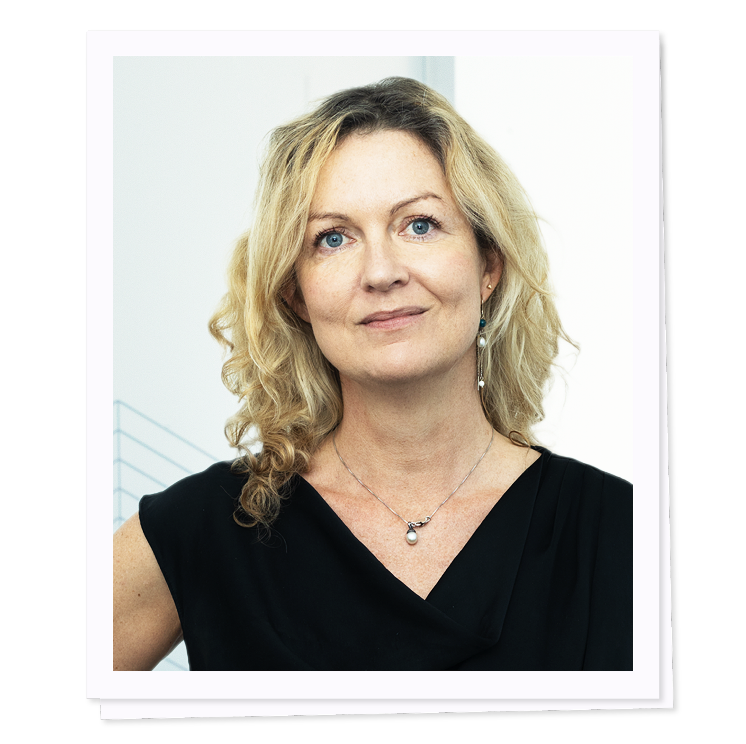 Portrait of a woman with blonde curly hair, wearing a black top and a pearl necklace, against a light background.