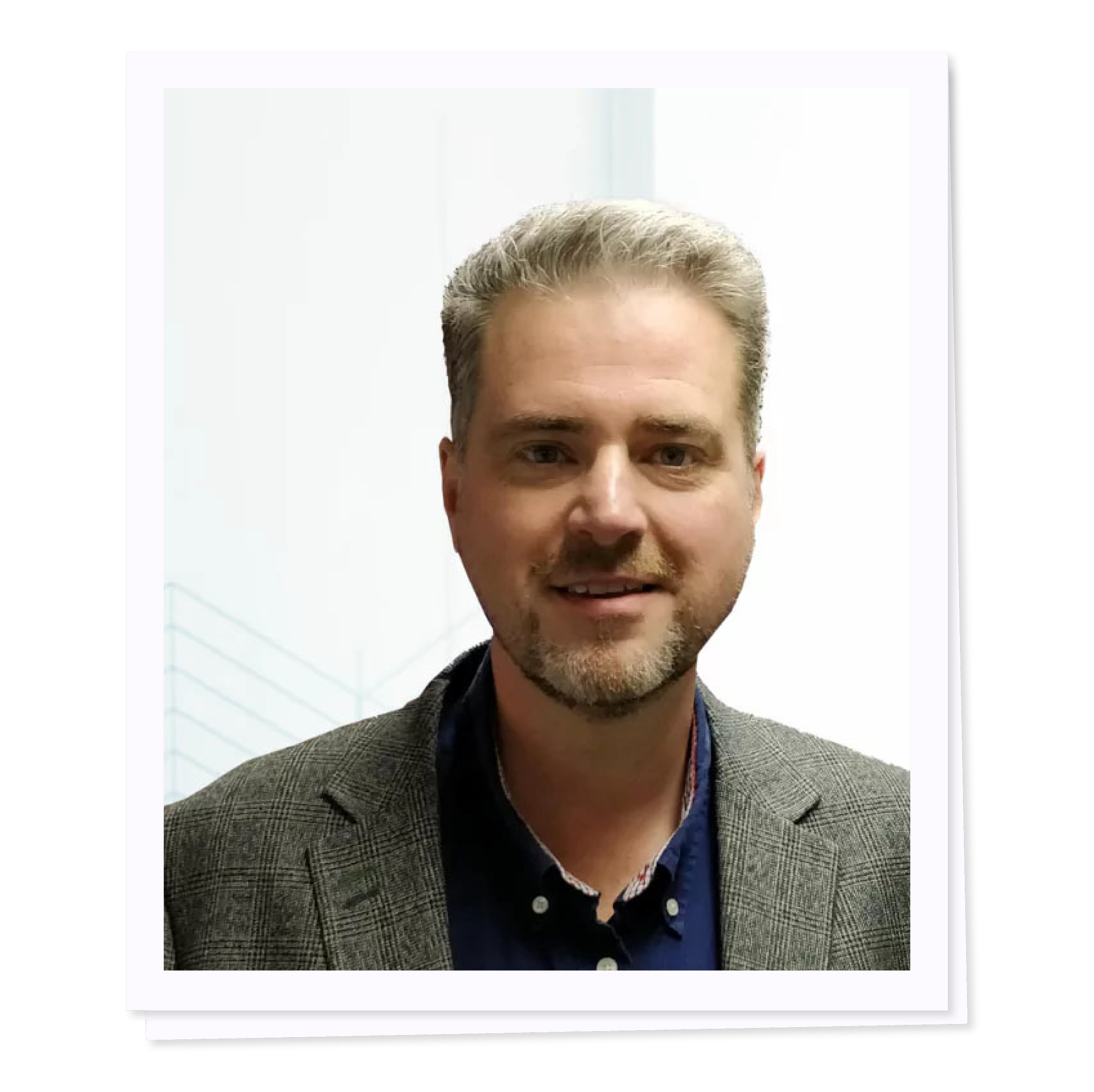 Portrait of a man with short gray hair, wearing a gray blazer and dark shirt, standing against a light background.